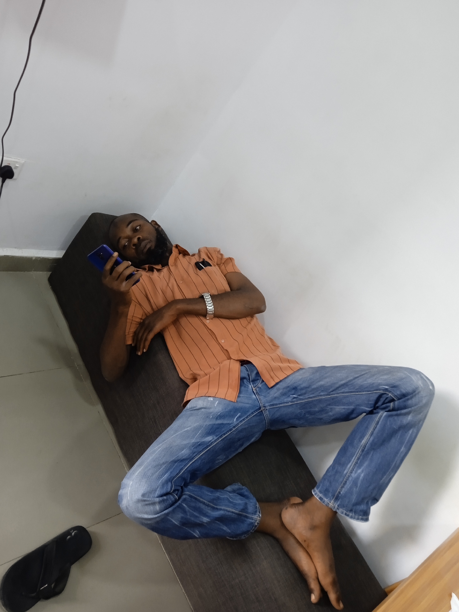 A man is lounging diagonally on a dark-colored sofa or bench indoors, with a white wall and white tiled floor in the background. He is holding a blue smartphone in his left hand and looking at it. He is wearing a short-sleeved, orange-striped shirt, a silver wristwatch, and blue jeans. He is barefoot. A black sandal is on the floor near the sofa. A wooden structure (possibly part of a cabinet or another piece of furniture) is visible near his feet. The location is presumed to be in Jos, Nigeria.