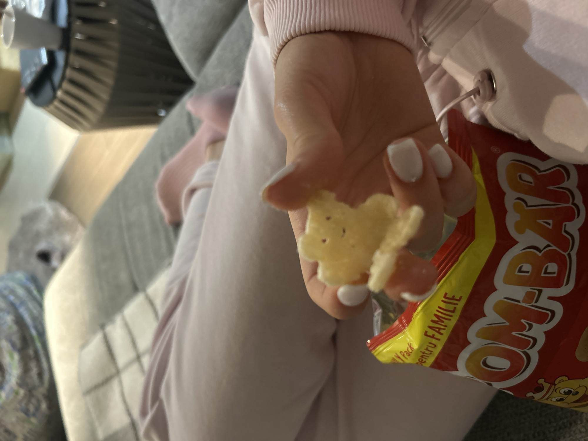 A close-up shot depicts a person holding an "OMB-BAR" chip in their hand. The person is wearing a pink sweatshirt and sweatpants. The background consists of a gray couch. The location of the photo is unknown.