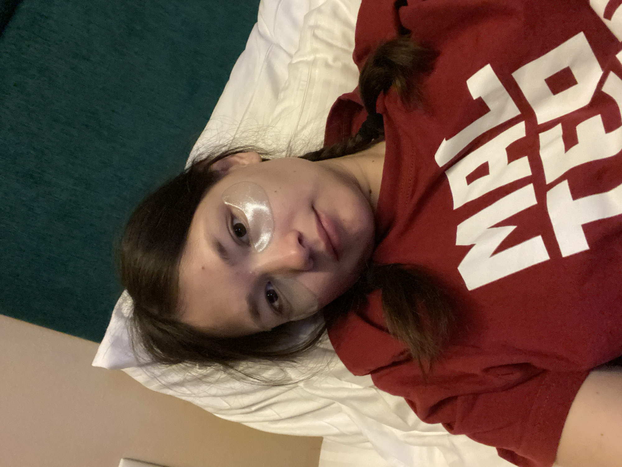 A young person with dark hair, styled in two pigtails, lies on their back in an indoor setting, likely a bedroom or hotel room in Peresvet, Russia. They are wearing a maroon or dark red t-shirt that features large white capital letters across the chest. The text "ANIMAL" is clearly visible on the shirt. The person has a neutral to slightly relaxed expression and is looking directly towards the camera. Two translucent, slightly shimmery under-eye patches are applied beneath their eyes, indicating a skincare routine or moment of relaxation.
The subject's head rests on a white pillow, and white bedding is visible around them. A dark green blanket or duvet occupies the upper left portion of the frame, contrasting with the lighter tones of the bedding. A light-colored wall or surface is also visible in the lower left background. The image is captured from a slightly elevated perspective, with the person positioned diagonally across the frame.