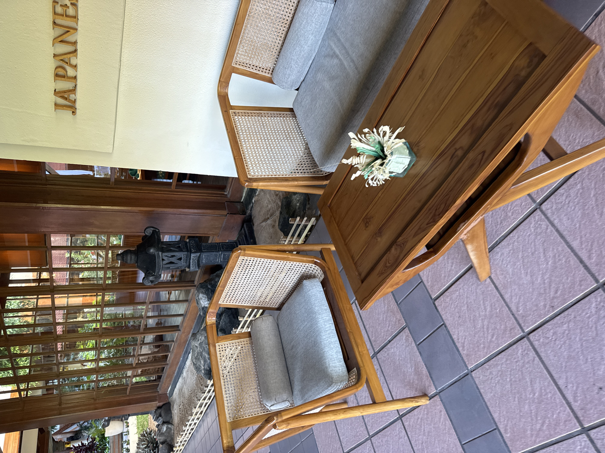 The photograph shows a seating area in Kuta Selatan, Indonesia, with a sofa and chairs arranged around a wooden table. A flower arrangement sits on the table. The area appears to be outdoors, possibly a patio, with a view of plants and a decorative lamp visible through a lattice screen. The ground is tiled. The sign "JAPANESE" is visible above, suggesting the location might be a Japanese restaurant or themed area.