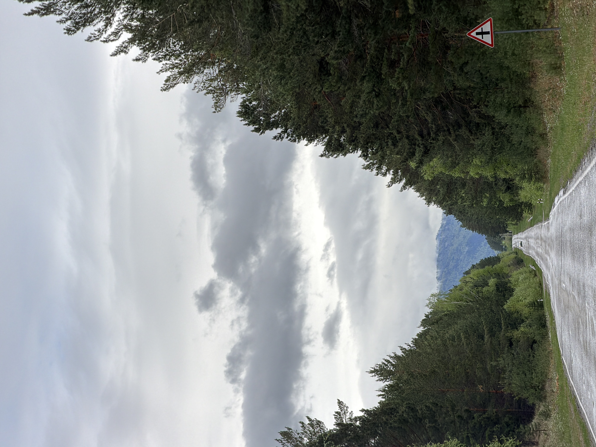 The image depicts a road in the vicinity of Сизая, Russia. The road is wet, likely due to recent rain, and lined with dense trees on both sides. A road sign warning of a crossroad is visible on the right side of the road. In the distance, mountains can be seen partially obscured by clouds. There are no people visible in the image, but a vehicle is seen further down the road. The sky is overcast with heavy, gray clouds.