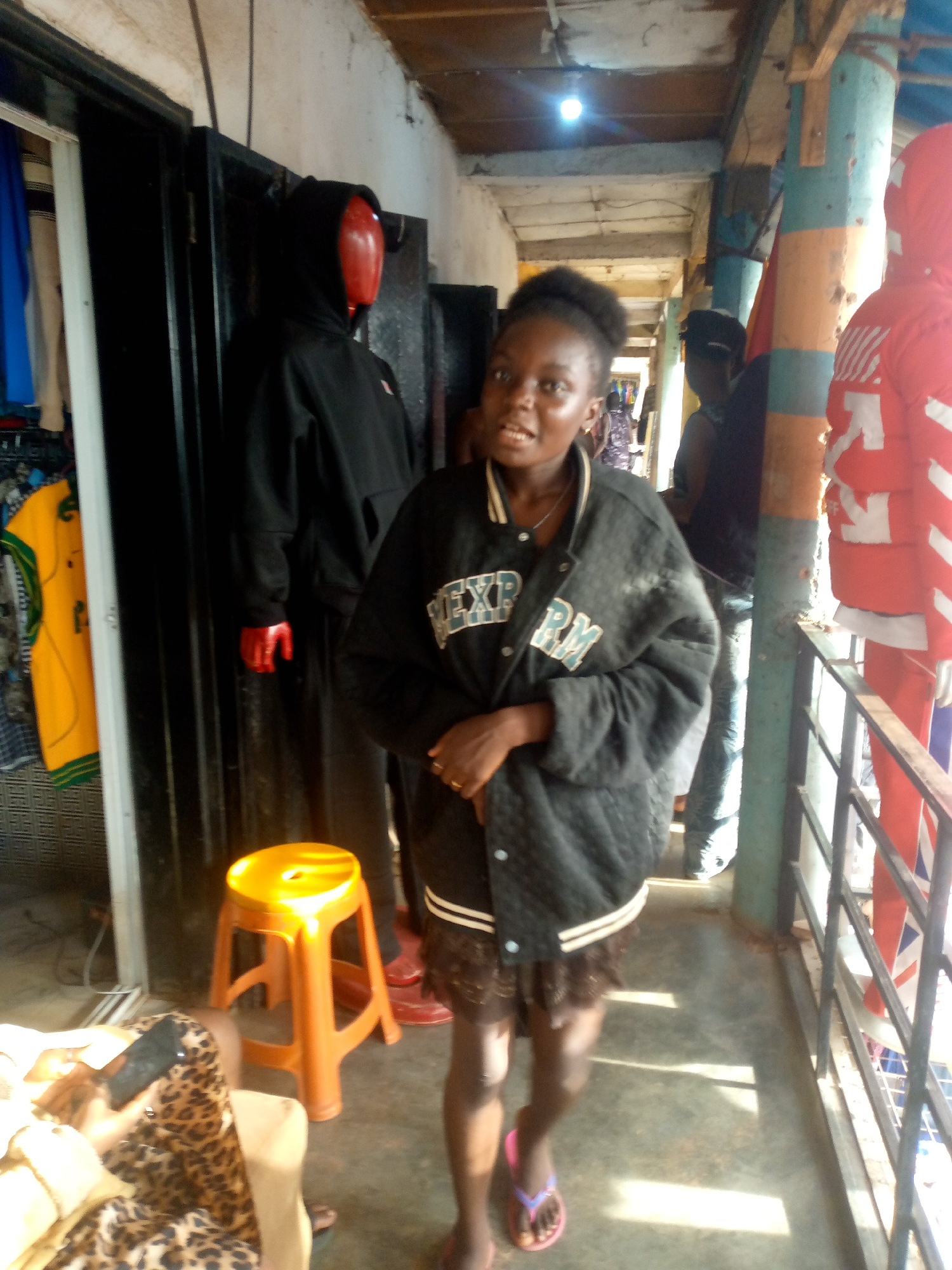 A young woman stands in the corridor of a Bukuru, Nigeria retail establishment. She is wearing a black varsity jacket and flip-flops. A mannequin in a black hoodie stands behind her and to the left, adjacent to an orange stool. Displayed clothing is visible on the left. Another mannequin clad in red is on the right, adjacent to a metal railing. Other people are visible in the corridor beyond her.