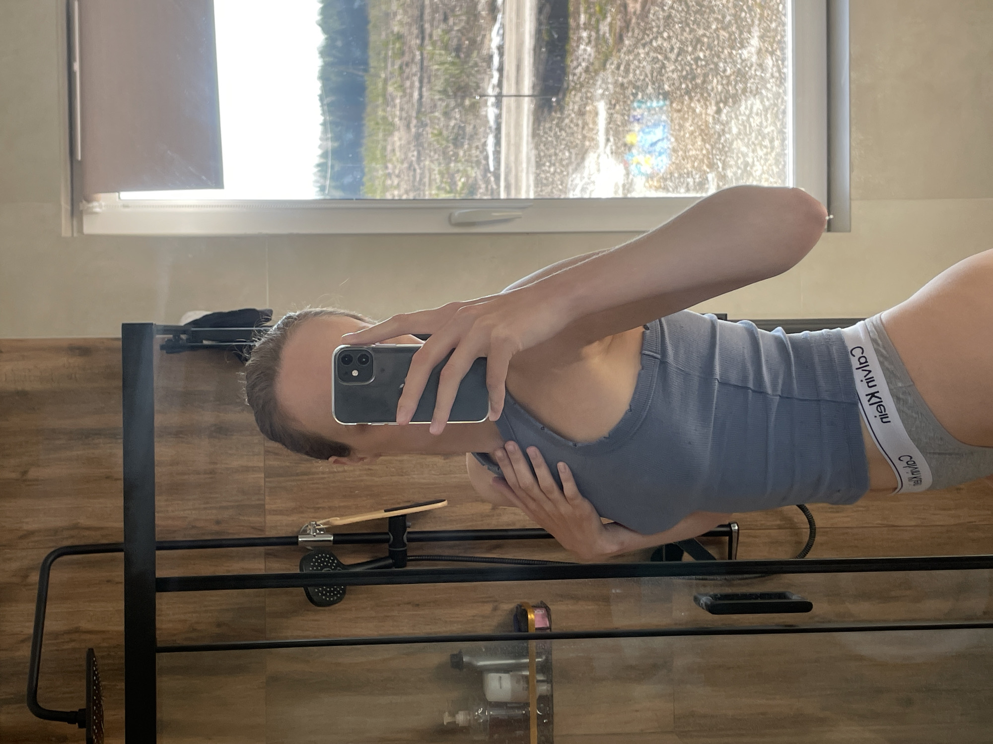 A person is taking a selfie in a bathroom. The person is wearing a gray tank top and Calvin Klein underwear. The bathroom has wood-look walls and a window with a view of trees and a field. A black shower caddy is visible in the foreground. The location is not discernible as Подшивалово, Russia, from the given image.
