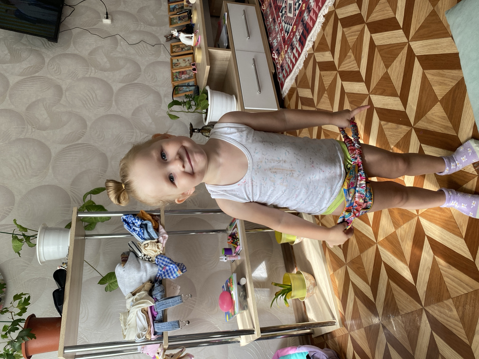 A young blonde girl with blue eyes and her hair in a bun is standing in what appears to be a living room in Otchor, Russia. She is wearing a white tank top and colorful shorts, with one sock on her left foot that is purple and adorned with small white flowers. The girl is pulling at the waistband of her shorts.
The room has a patterned wallpaper and a wooden floor with an intricate geometric design. To the left of the girl is a tall, multi-tiered shelving unit holding various clothing items, shoes, and potted plants. Behind the shelving unit, a television is mounted on the wall. To the right of the girl is a wooden cabinet with two drawers, and next to that, a colorful rug with a fringe. The lighting suggests it is daytime, possibly late morning or afternoon. The overall scene depicts a casual domestic setting. There is no visible text in the image.