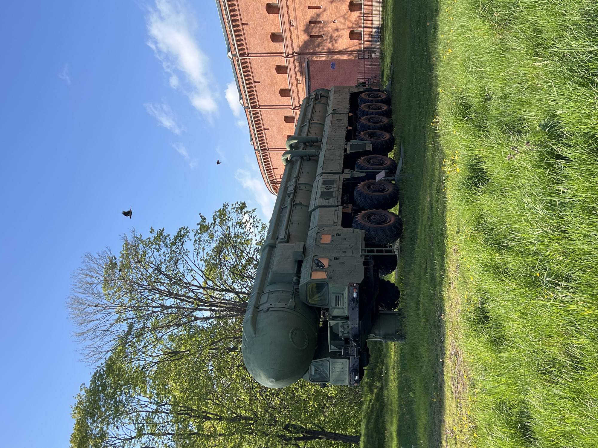 The image shows a TEL (Transporter Erector Launcher) vehicle parked on a grassy field in Saint Petersburg, Russia. The vehicle, likely a ballistic missile launcher, is green and has multiple axles with large tires. A red brick building is visible in the background. Two birds are flying in the blue sky with scattered white clouds. A deciduous tree is in the foreground.