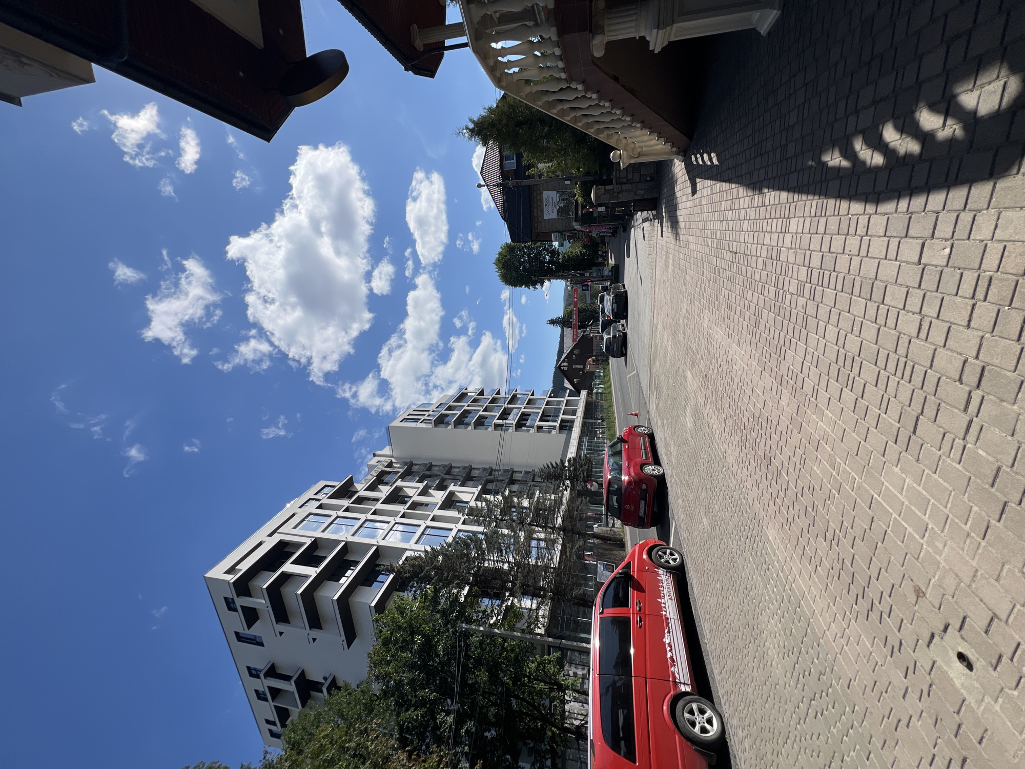 The image depicts a street scene in Yaremche, Ukraine, under a clear blue sky with scattered white clouds. A modern, multi-story building with white facade and dark-framed balconies dominates the left side of the frame, with mature trees lining its base. A red van and a smaller red car are parked on the paved street. To the right, a stone staircase with ornate white balustrades leads up to a building. The street is paved with reddish-brown bricks, and shadows from the surrounding structures fall across it. In the background, more residential buildings and trees are visible, with a mountainous landscape suggested in the distance. The overall impression is a sunny day in a well-maintained urban or semi-urban environment.