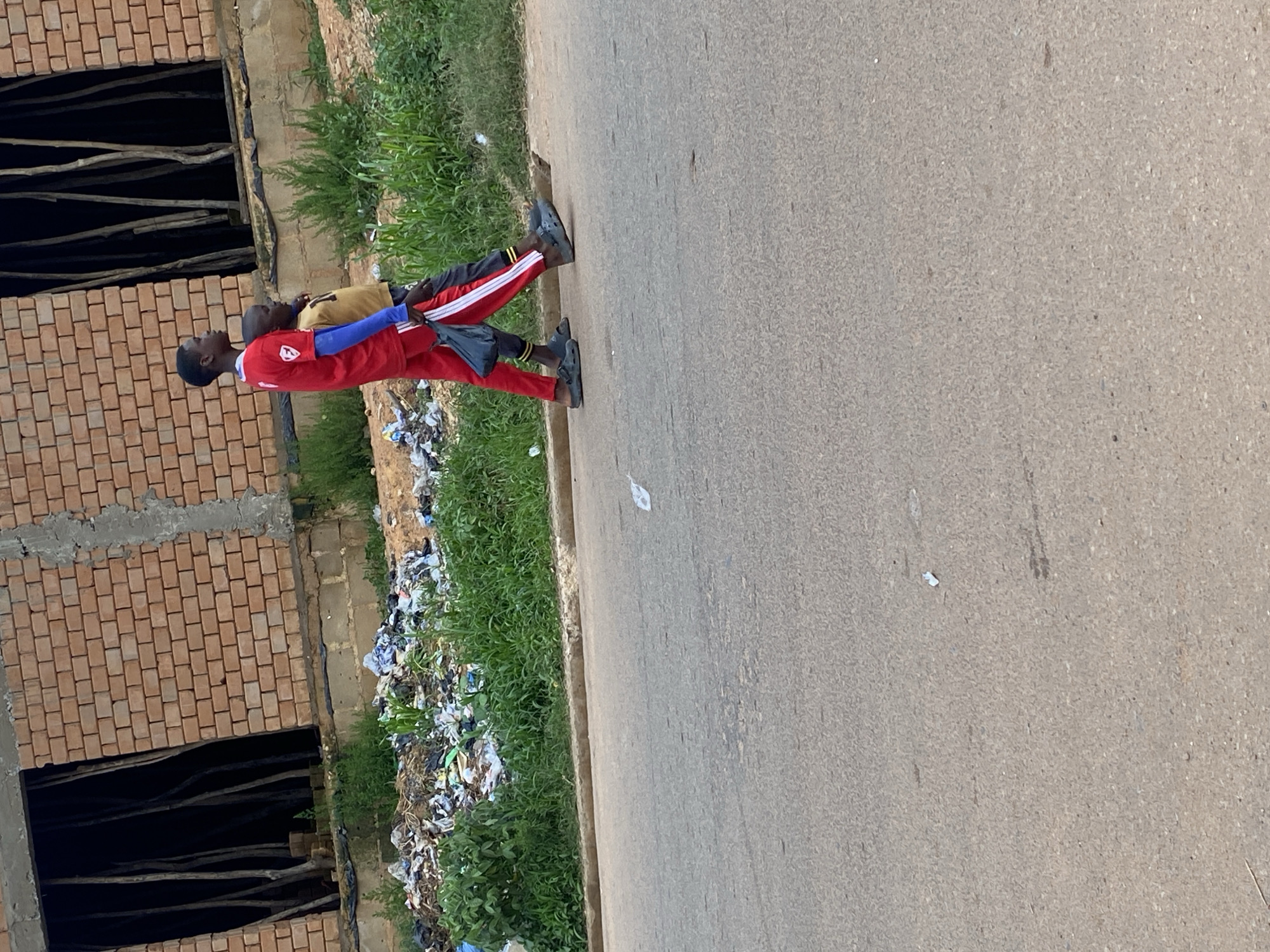 Two young males, one in a red shirt and red pants with white stripes, and the other in a yellow shirt, walk along a paved road in Bukuru, Nigeria. The male in red is carrying a black plastic bag. Behind them, a construction site with brick walls and open window frames is visible, with debris and overgrown grass along the roadside.