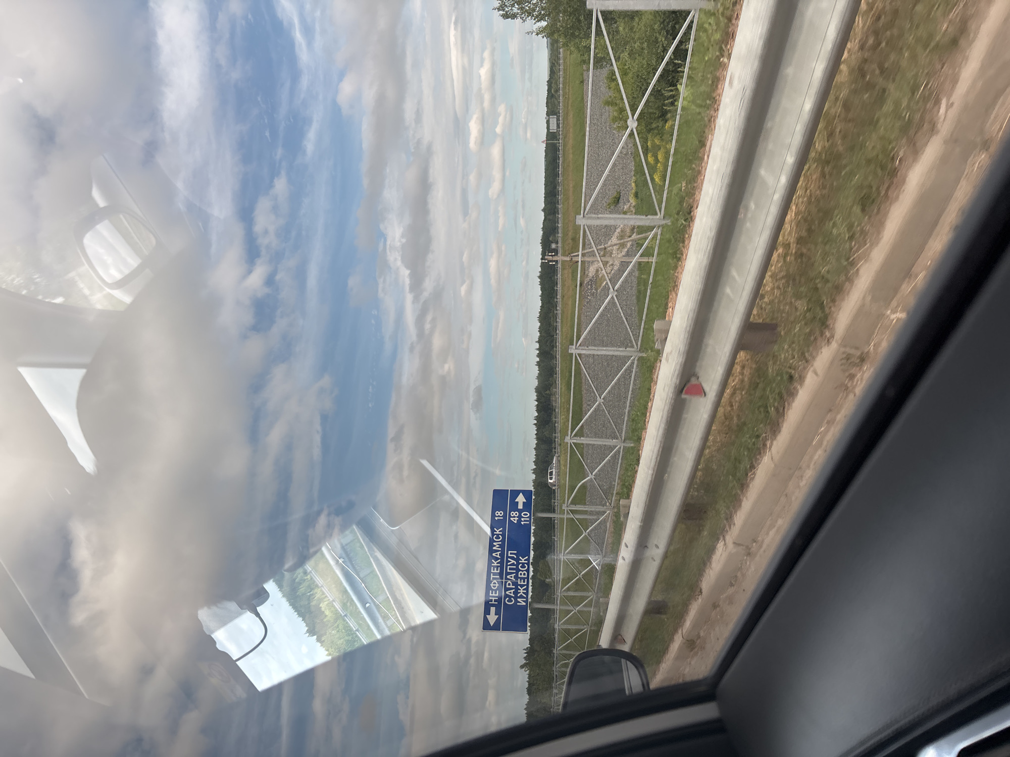 The image shows a view from inside a vehicle, looking out a window. In the foreground, the interior of the car is visible, including a dark-colored dashboard and a portion of the side window frame. Reflected in the window are the sky with clouds and some interior elements of the car. Outside, a blue road sign with white lettering is visible. The sign indicates directions to "НЕФТЕКАМСК 18" (Neftekamsk 18) to the left, and "САРАПУЛ 48" (Sarapul 48) and "ИЖЕВСК 110" (Izhevsk 110) to the right. A white metal guardrail runs alongside the road. Beyond the guardrail, there is a grassy area with some yellow wildflowers, followed by a line of trees forming a dense forest. A white passenger car is visible in the distance on the road. The sky is partly cloudy with patches of blue. The scene appears to be a highway location in Russia, likely on the outskirts of Kambarka.