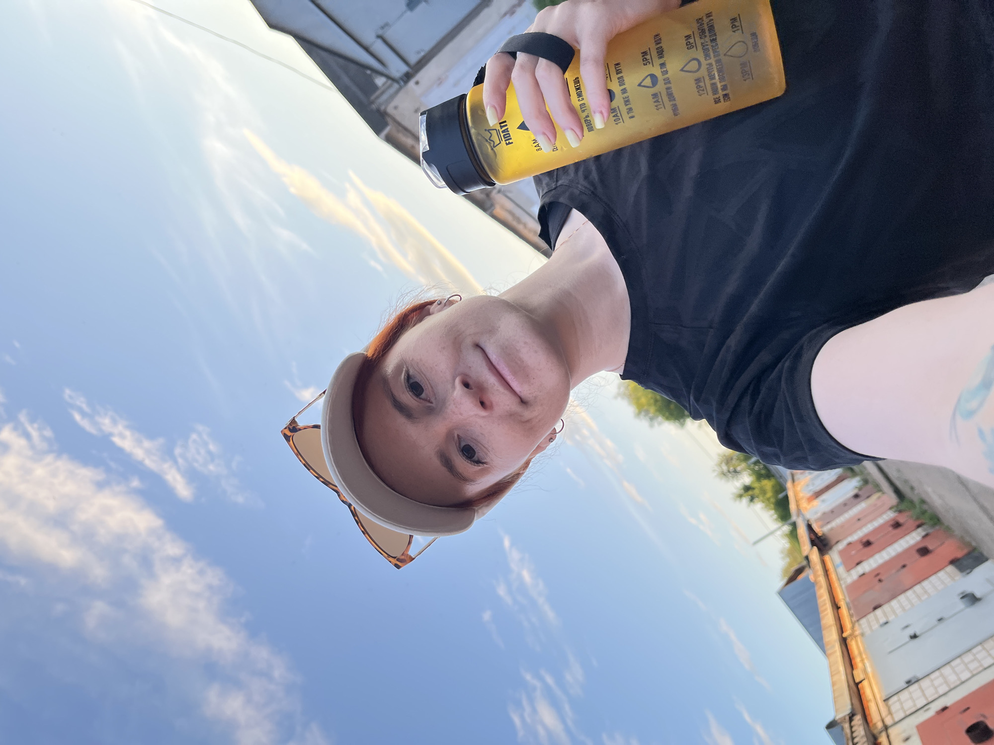 A fair-skinned female subject with reddish hair and visible freckles is depicted in an outdoor selfie. She wears a beige baseball cap with tortoise-shell framed sunglasses pushed onto the cap and a black short-sleeved t-shirt. Small silver hoop earrings are visible on her ears. Her right hand firmly grips a translucent yellow hydration bottle, featuring a black lid and wrist strap. The bottle displays the brand name "FIDATI" with a crown logo, along with time markers from 8 AM to 6 PM and motivational text in Russian. A tattoo is partially visible on her left arm. The background features a clear blue sky with scattered stratocumulus clouds and a prominent sun glare emanating from the upper right quadrant. Below the horizon line, a linear arrangement of single-story structures resembling garages or storage units is visible, characterized by doors of varying colors including red, white, and gray. Green foliage from trees is present above and partially between some of these structures. The scene is located in the city of Chaikovsky, Russia.