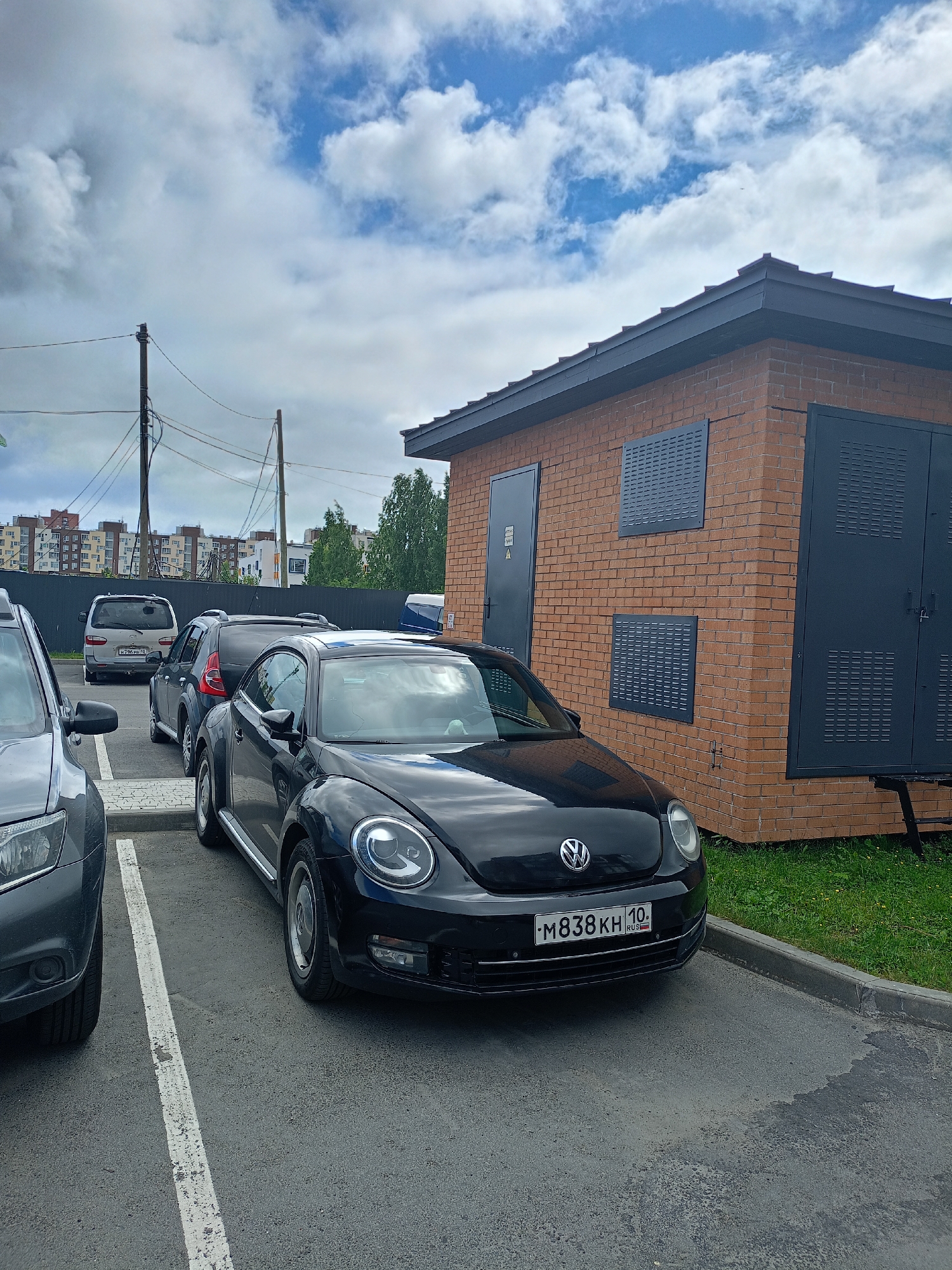 A black Volkswagen Beetle is parked in a paved lot next to a brick building. The building has a dark gray roof and dark gray double doors on the right side, and a single dark gray door on the left side. There are two dark gray rectangular grates on the brick wall between the doors. The car has a license plate that reads "M838KH 10". In the background, there are several other cars parked, a tall fence, apartment buildings, trees, and a cloudy sky. The car is positioned in the foreground, angled slightly towards the right. The scene appears to be in an urban or suburban area.