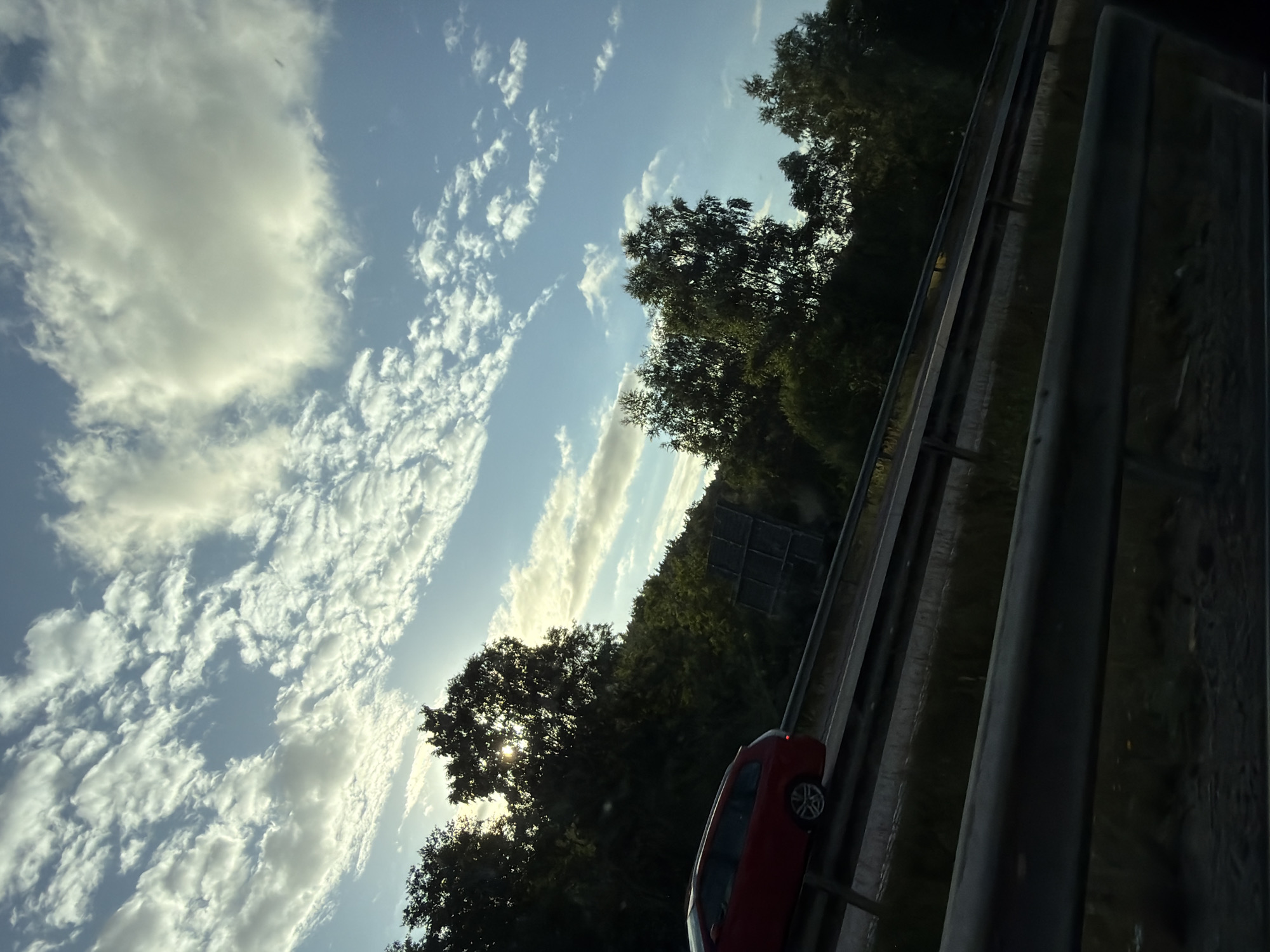 A red station wagon is seen driving on a highway in Kungsbacka, Sweden. The vehicle is in the left lane, moving away from the viewer. The highway is bordered by a guardrail and a dense line of trees and vegetation on the right. The sky above is a clear blue with scattered white, wispy clouds, illuminated by the sun which is partially obscured by the trees. The sun's rays are creating a lens flare effect visible through the foliage. The overall scene suggests daytime travel on a road surrounded by nature.