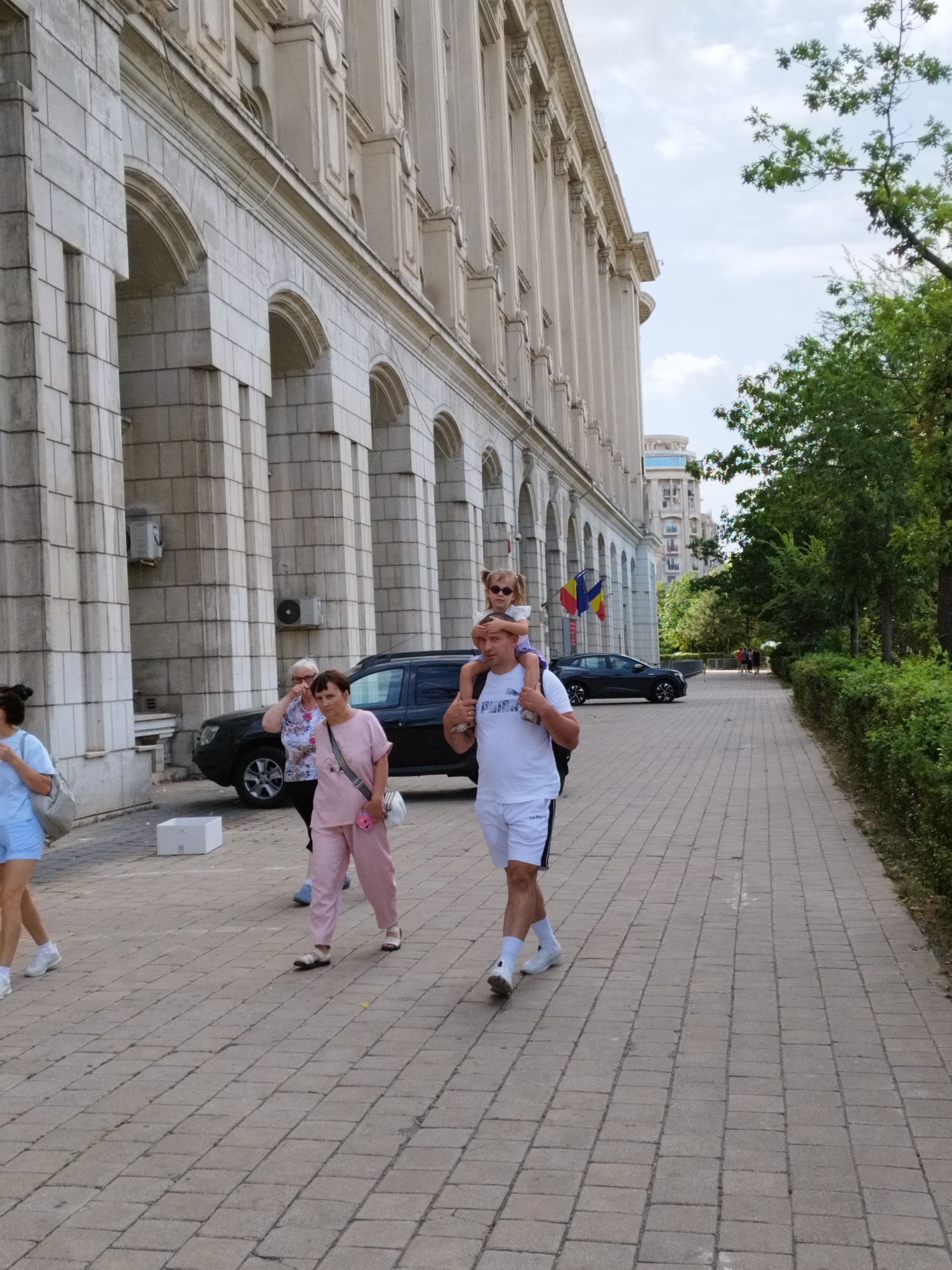 In Bucharest, Romania, a group of people walks along a paved sidewalk adjacent to a grand, classical-style building with arched colonnades. A man is carrying a young girl on his shoulders. The girl is wearing sunglasses and a white t-shirt. The man is wearing a white t-shirt and white shorts with black stripes. Two women are walking behind them, one in a pink tracksuit and the other in a white and floral patterned top and dark pants. A woman in a light blue outfit is walking further ahead on the left. A black car is parked behind the two women. In the background, trees and other buildings are visible. The sky is partly cloudy.