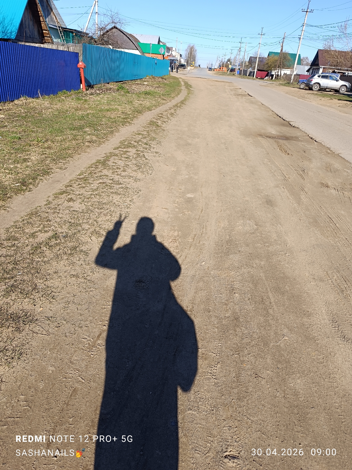 The image depicts a dirt road in the city of Chaikovsky, Russia. The road extends into the distance, lined with fences, utility poles, and houses. A shadow of a person, possibly taking the picture, is cast onto the road. The photo was captured on April 30, 2026, at 9:00 AM using a Redmi Note 12 Pro+ 5G.