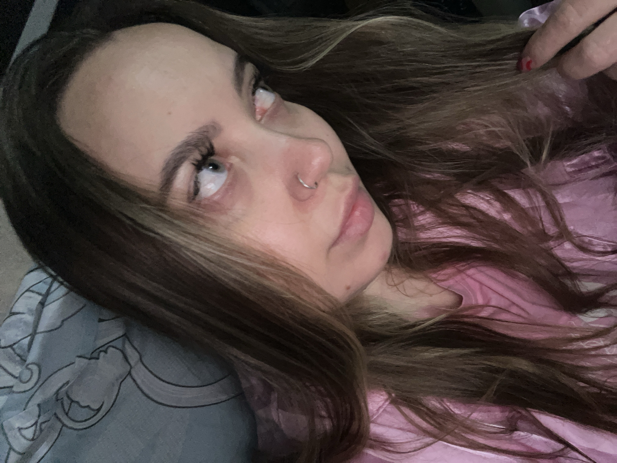 A woman lies on a pillow, wearing a pink top. She has long brown hair with blonde highlights and a nose ring. Her gaze is directed upwards and to the left. The background is out of focus. There is no information to determine if this image was taken in Novopokrovskaya, Russia.