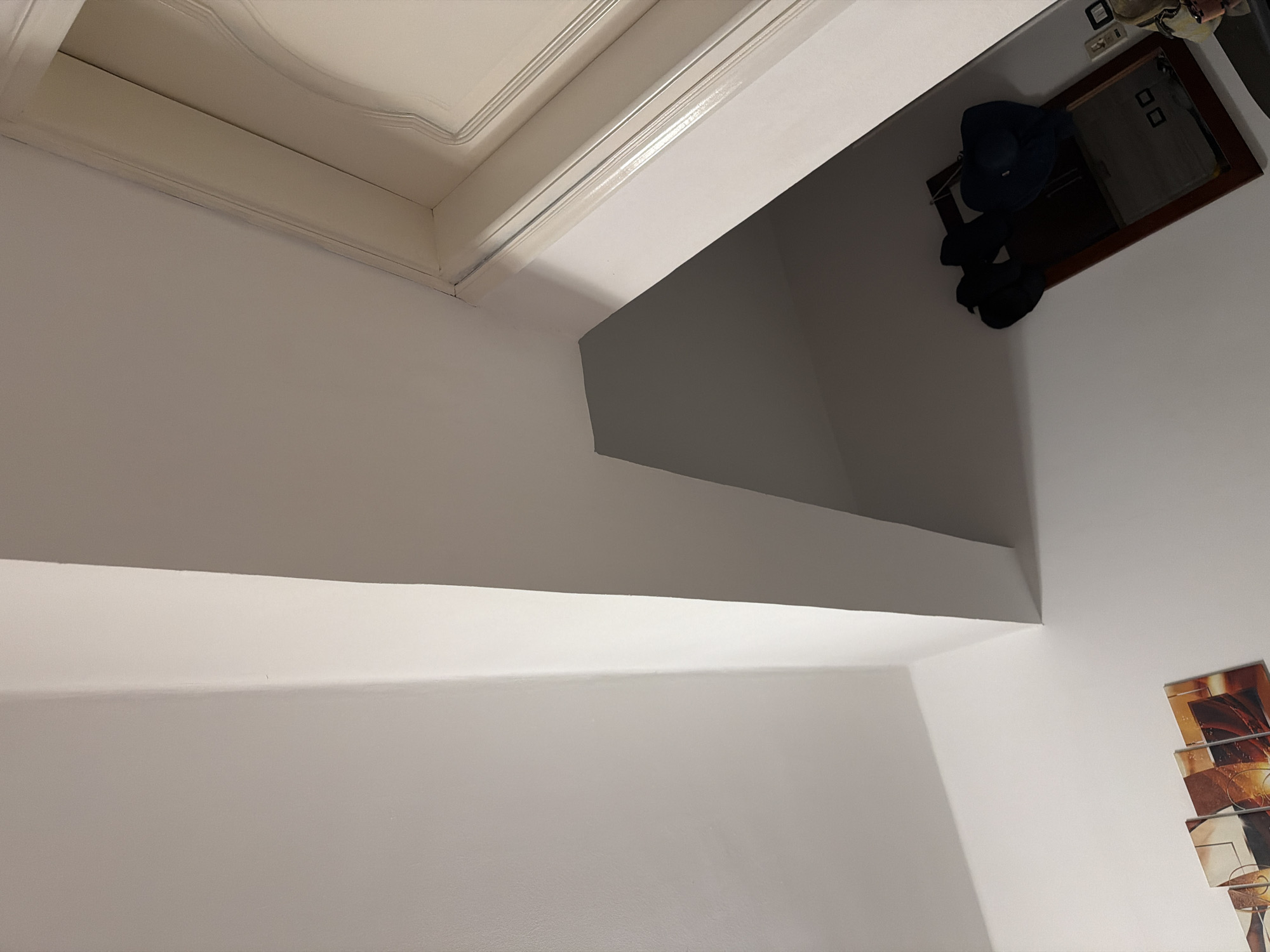 The image depicts an interior scene, likely a hallway or entryway. The dominant features are a white wall and ceiling, with architectural lines suggesting a corner or an angled structure. To the right, a portion of a white door frame with decorative molding is visible, suggesting a doorway. Lower down, a dark-colored mirror with a wooden frame hangs on the wall, adorned with several dark hats. In the bottom left corner, a section of a multi-panel artwork with abstract, warm-toned designs is partially visible. The lighting appears to be dim, creating shadows and highlighting the textures of the surfaces. The scene does not provide any specific visual cues that would link it to Sharm El Sheikh, Egypt.