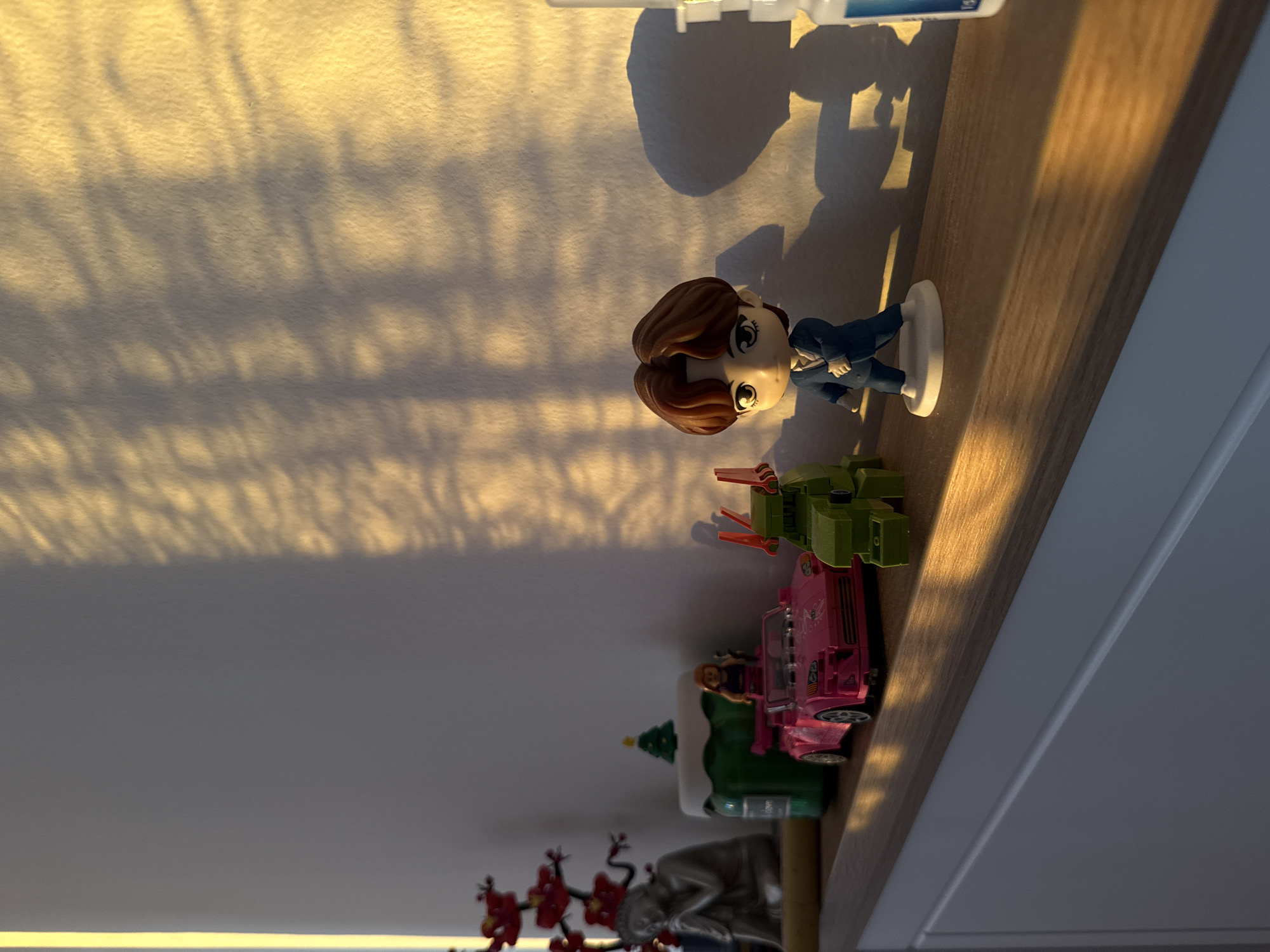 A shelf in a room is shown from an upward angle. On the shelf are various toys and figurines. Starting from the bottom and going up, there is a statue of a person with red cherry blossom-like decorations. Above that is a green Christmas tree decoration, followed by a pink toy car with a figurine inside. Next to the car is a green toy vehicle. Above these, there is a figurine of a person wearing a blue suit and standing on a white base. To the right, there is also a white container. Light from a window is casting long shadows across the wall behind the shelf. The location in Kragujevac, Serbia is not able to be determined based on the objects in the image.