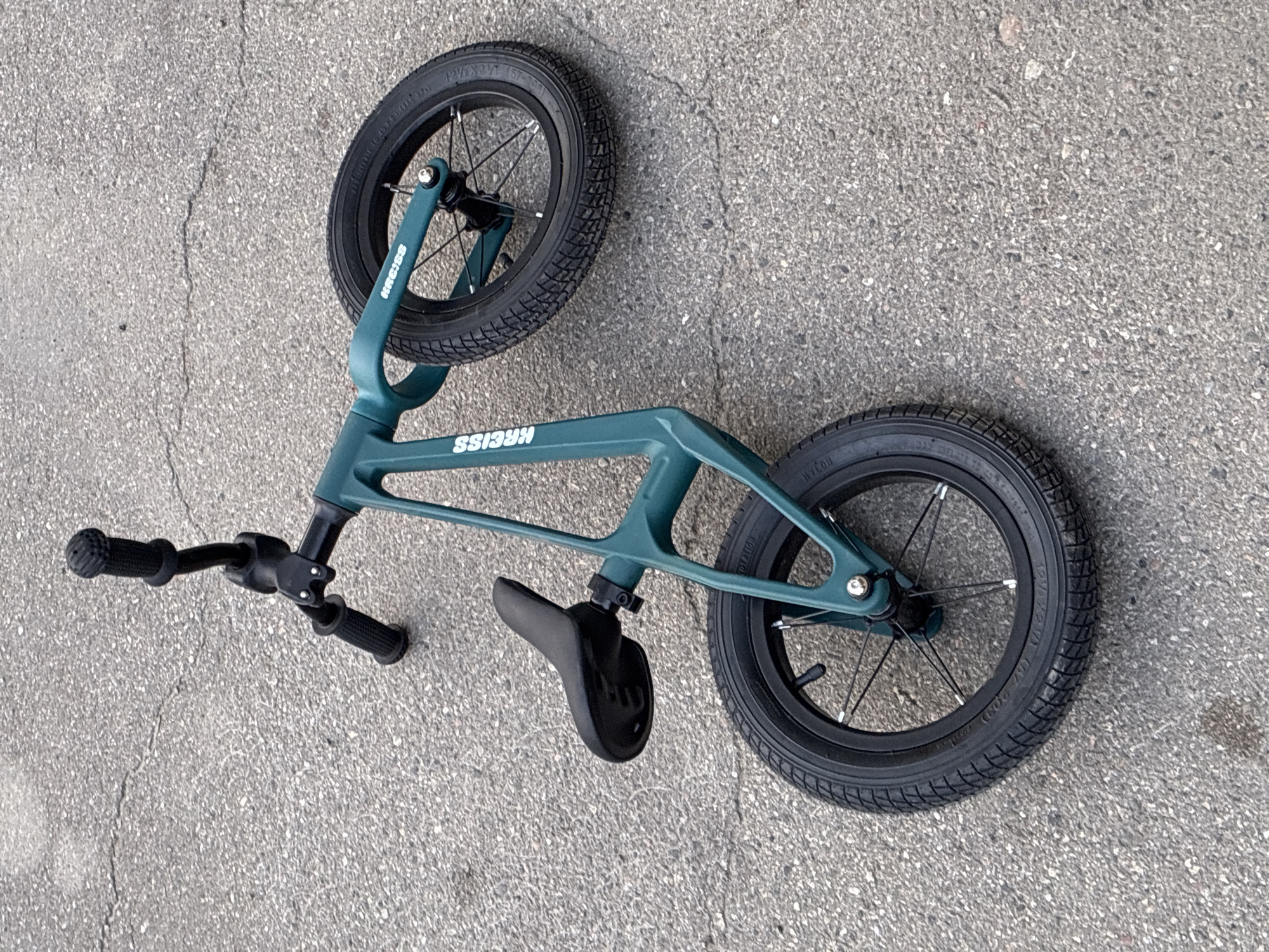 A teal balance bike is lying on its side on cracked asphalt. The bike has black handlebars with grips, a black seat, and two black wheels with thin spokes. The brand name "KRISS" is visible in white lettering on the frame of the bike. The asphalt appears to be a road or pathway, and there is no other visible debris or context in the image. The scene is outdoors. The location in the city of Markova, Russia is not discernible from the image.