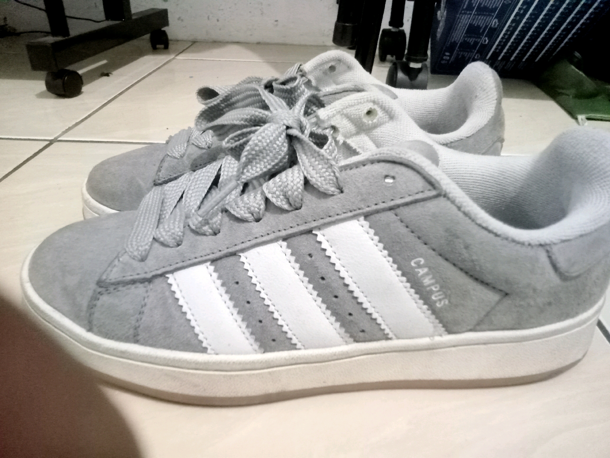 Two grey Adidas Campus sneakers are shown from a slightly elevated angle, resting on a light-colored tiled floor. The shoe in the foreground is a suede material with white Adidas stripes on the side and the word "CAMPUS" printed in white above the stripes. The laces are grey and tied in a bow. The other shoe, slightly behind and to the left, is similar in style and color, also with grey laces. The background includes a dark-colored object with wheels, possibly a cart or stand, and a dark blue patterned box. The lighting is dim, casting shadows on the floor.