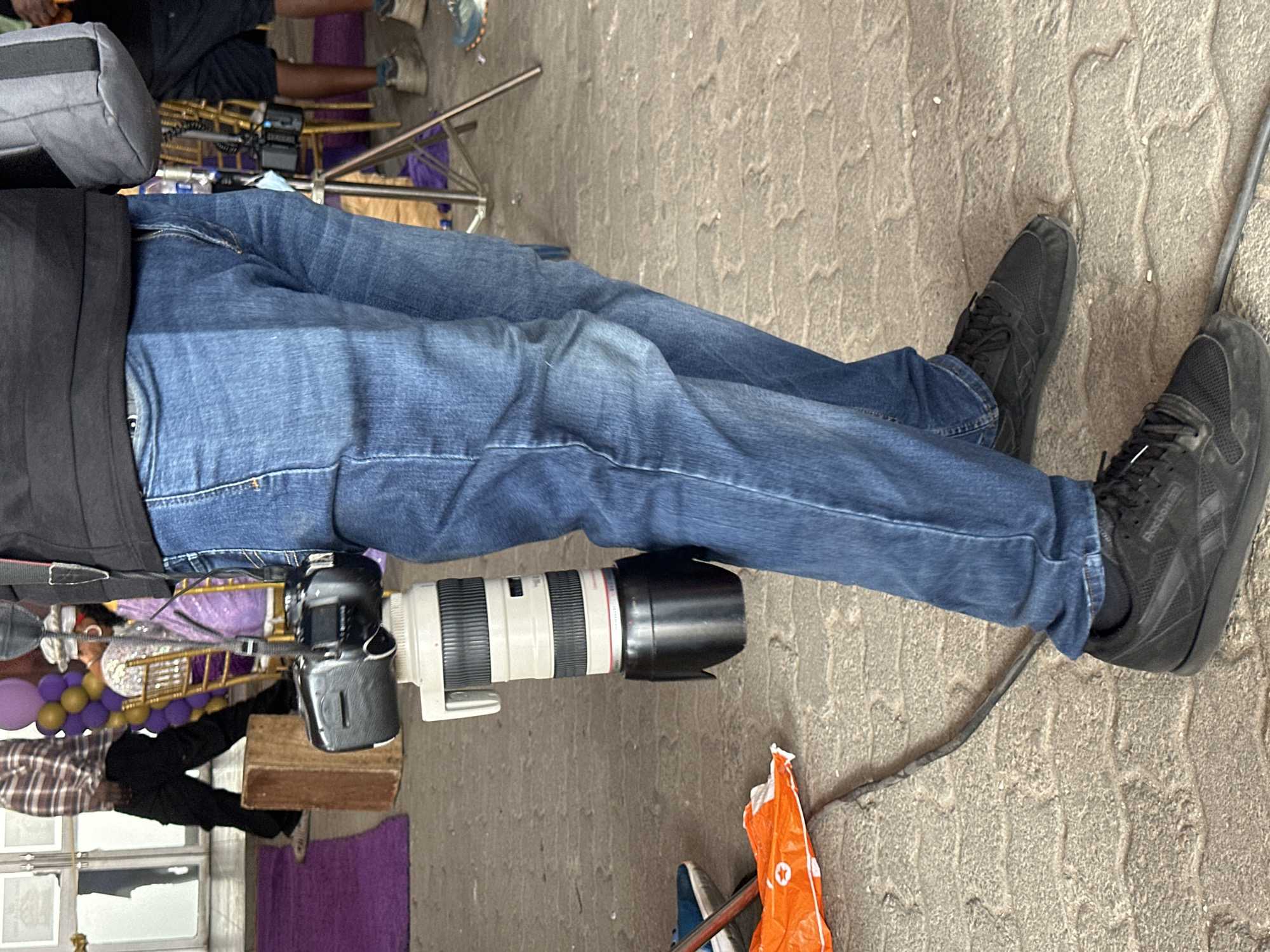 The image shows a person wearing blue jeans and black sneakers, holding a camera with a large lens. A second person is partially visible in the background. There are balloons and a decorated backdrop suggesting a possible event setup, but the exact type of event is not clear. The setting appears to be outdoors on a paved surface. It is not possible to determine the location as Lagos, Nigeria with the provided details.