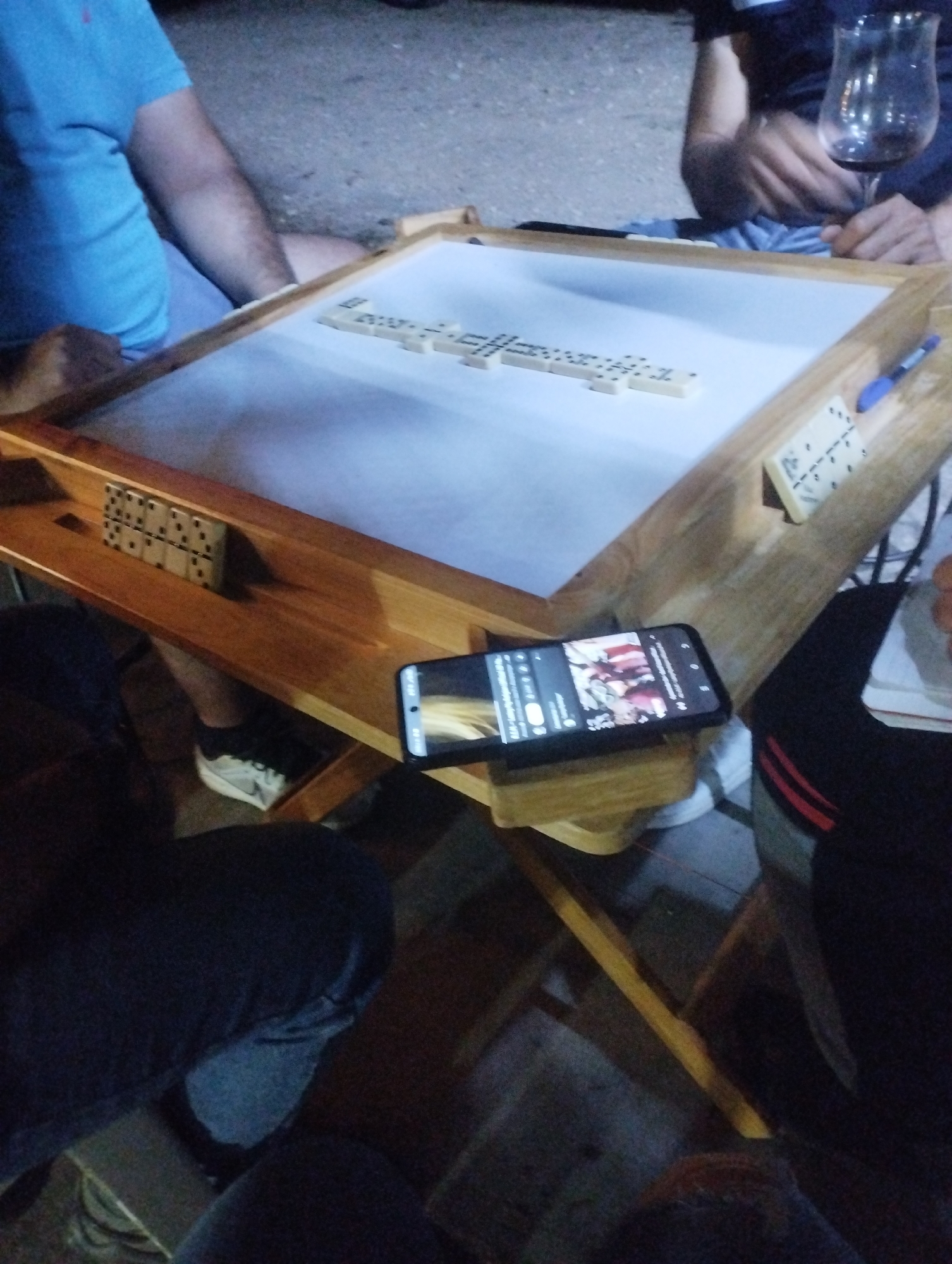 The scene depicts a group playing dominoes in Tinaquillo, Venezuela. A wooden domino table with a white playing surface is the focal point. Several dominoes are in play on the white surface, with additional pieces held in wooden racks along the table's edges. A black smartphone sits on the table's edge, displaying a YouTube video. Three people are visible around the table: one wearing a blue shirt, another with a dark shirt and holding a wine glass, and a third wearing pants with a red stripe. The environment suggests an outdoor setting, possibly an evening gathering.