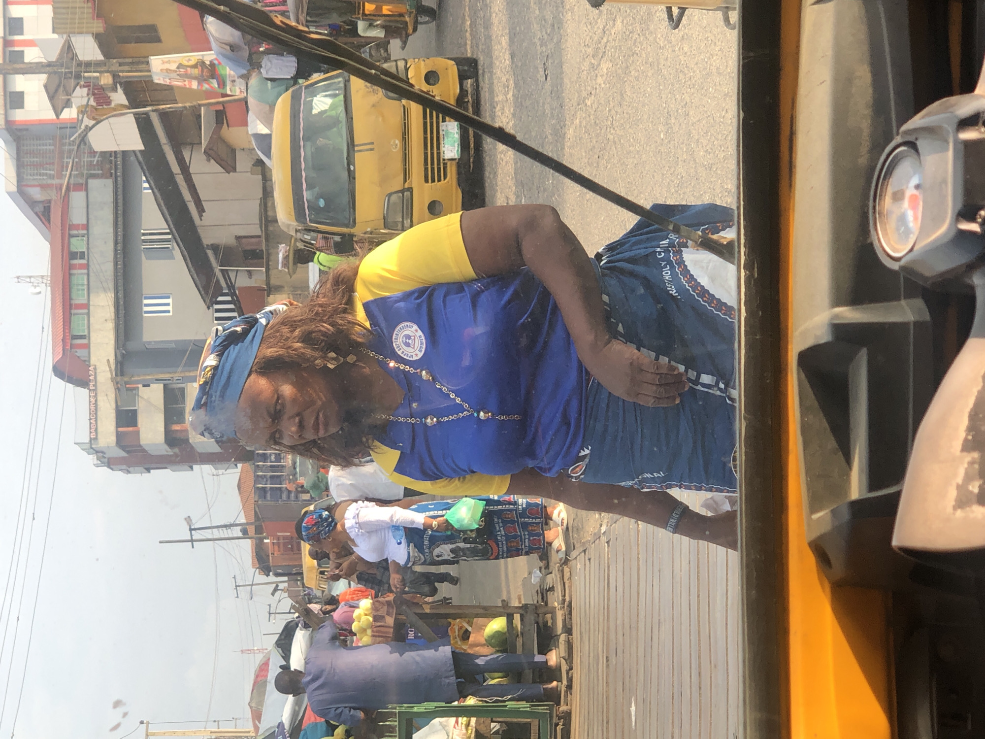 The image depicts a street scene in Lagos, Nigeria. In the foreground, a woman wearing a blue and yellow shirt and a blue patterned skirt is looking to the left. She has a blue headscarf and is wearing earrings and a necklace. Behind her, a yellow minibus is visible. The background shows a bustling market environment with various people, stalls, and buildings. A man in a blue outfit is seen to the left, and another woman in a white top and blue skirt is standing near a fruit stall. Power lines and poles are visible above the buildings. The overall scene is one of a lively urban marketplace.