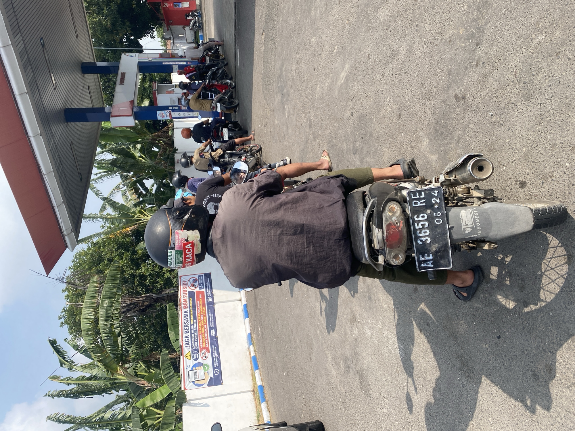 The image depicts a sunny outdoor scene at a Pertamina gas station in Maospati, Indonesia. A line of motorcycles is waiting to refuel. The foreground features a person on a motorcycle wearing a black helmet and a dark grey long-sleeved shirt, with their back to the camera. The motorcycle's license plate reads "AE 3656 RE" and "06 24". In the background, several other motorcycles and riders are visible, queuing under a gas station canopy. A red structure with "PERTAMINA" signage is present, along with gas pumps. A banner with Indonesian text and graphics about fuel subsidies is displayed on a white wall. Banana trees and other foliage are visible in the background. The ground is paved asphalt.