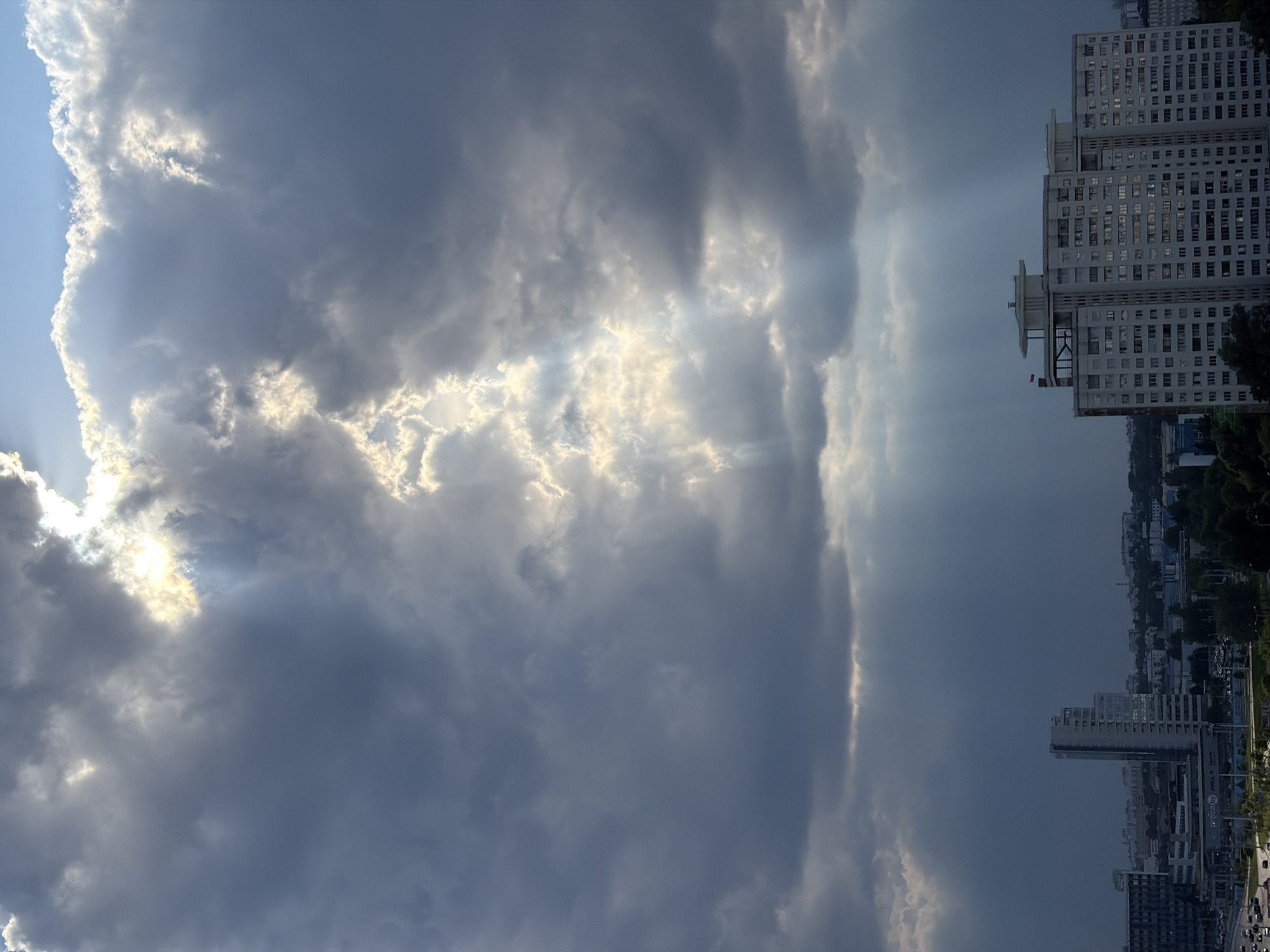 The image captures a dramatic sky over a city landscape, likely in or near Бараўлянскі сельскі Савет, Belarus. The sky is dominated by dark, heavy clouds, but a powerful beam of sunlight pierces through them, creating a striking contrast and illuminating a section of the clouds with a bright, golden glow. These sunbeams extend downwards, suggesting an impending or recent rain shower.
In the lower portion of the image, the urban environment is visible. A prominent, multi-story, light-colored apartment building with numerous windows is a central feature. To its left, another, darker building stands, appearing more modern and rectangular. Beyond these main structures, a dense arrangement of other buildings, some taller and others shorter, stretches across the horizon, interspersed with patches of green trees, indicating a residential or mixed-use urban area. The overall impression is a cityscape viewed from an elevated perspective under a dynamic and atmospheric sky.
