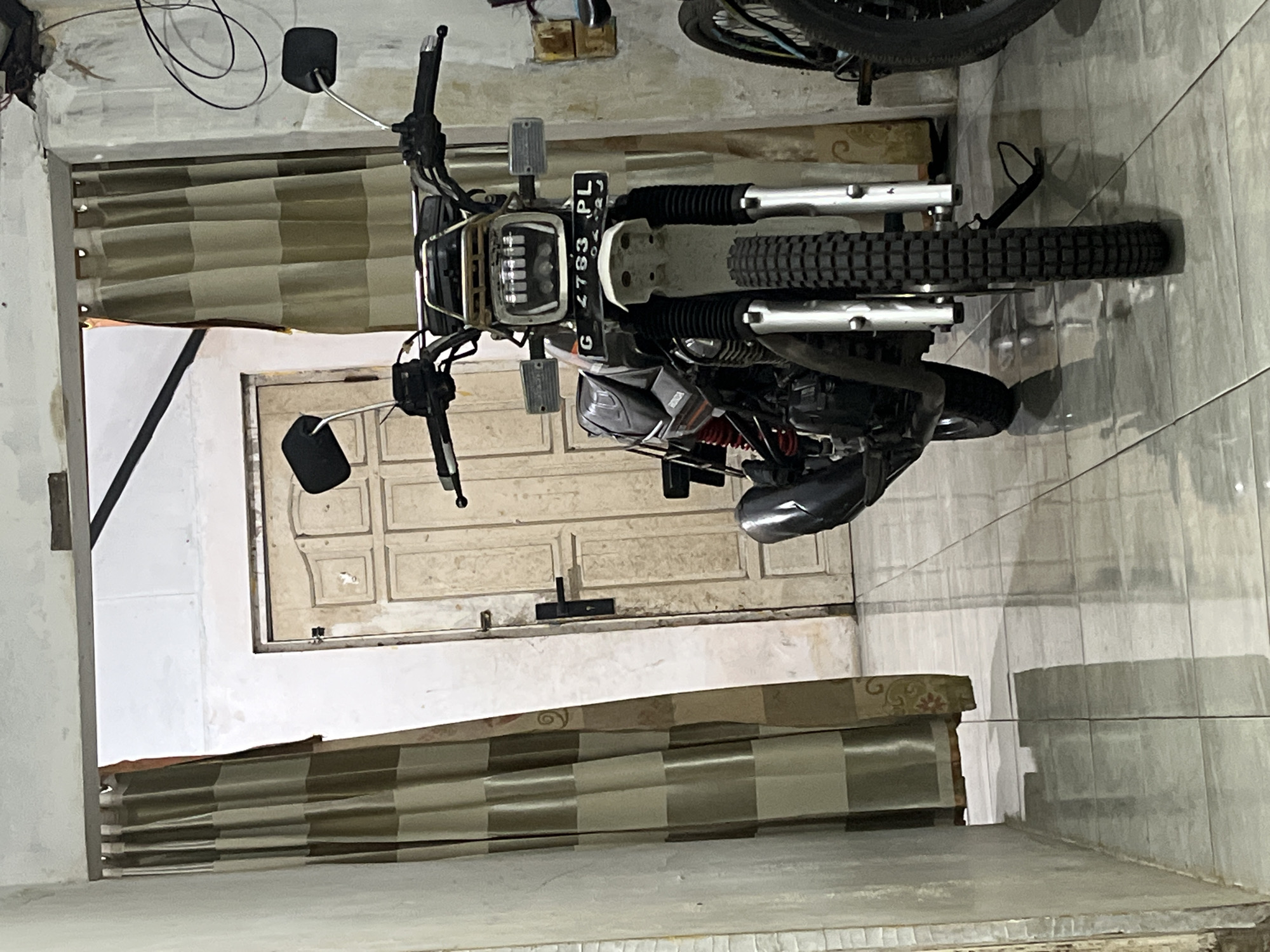 A motorcycle is parked indoors, facing forward, positioned centrally in the frame. The motorcycle is a dark color with silver accents on the handlebars, front forks, and a rack above the headlight. It has a black license plate on the front that reads "G 2763 PL". The motorcycle has dual mirrors on the handlebars and a prominent headlight assembly. The interior space appears to be a garage or workshop. To the left and right of the motorcycle are tan and brown striped curtains hanging from a doorway. In the background, there is a light-colored wooden door with panels. The floor is tiled with a reflective surface. The walls are a light color, with some visible discoloration and texture. A portion of another bicycle is visible on the right side of the frame, behind the motorcycle. The overall lighting suggests an indoor environment with artificial light. The presence of the motorcycle and bicycle suggests a residential or storage area within Pekalongan, Indonesia, possibly a home garage.