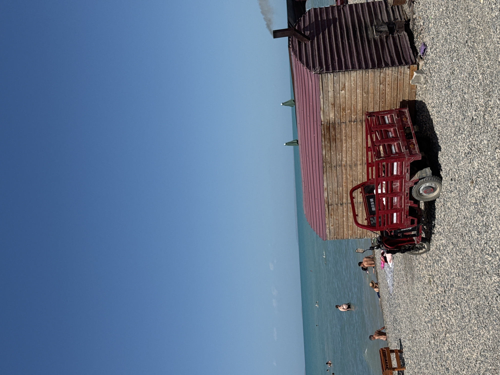 The image depicts a sunny day at a pebble beach in Zharbulak, Kazakhstan. The vast expanse of a turquoise sea stretches to the horizon under a clear blue sky. In the foreground, a small, red motorized tricycle is parked next to a rustic wooden structure with a corrugated red roof. Smoke billows from a chimney on the roof of the structure, indicating some activity within. Several people are visible in the shallow waters of the sea, some swimming and others relaxing. Scattered on the beach are a few beach chairs and umbrellas, suggesting a recreational area. The overall atmosphere is one of leisure and a pleasant summer day by the sea.