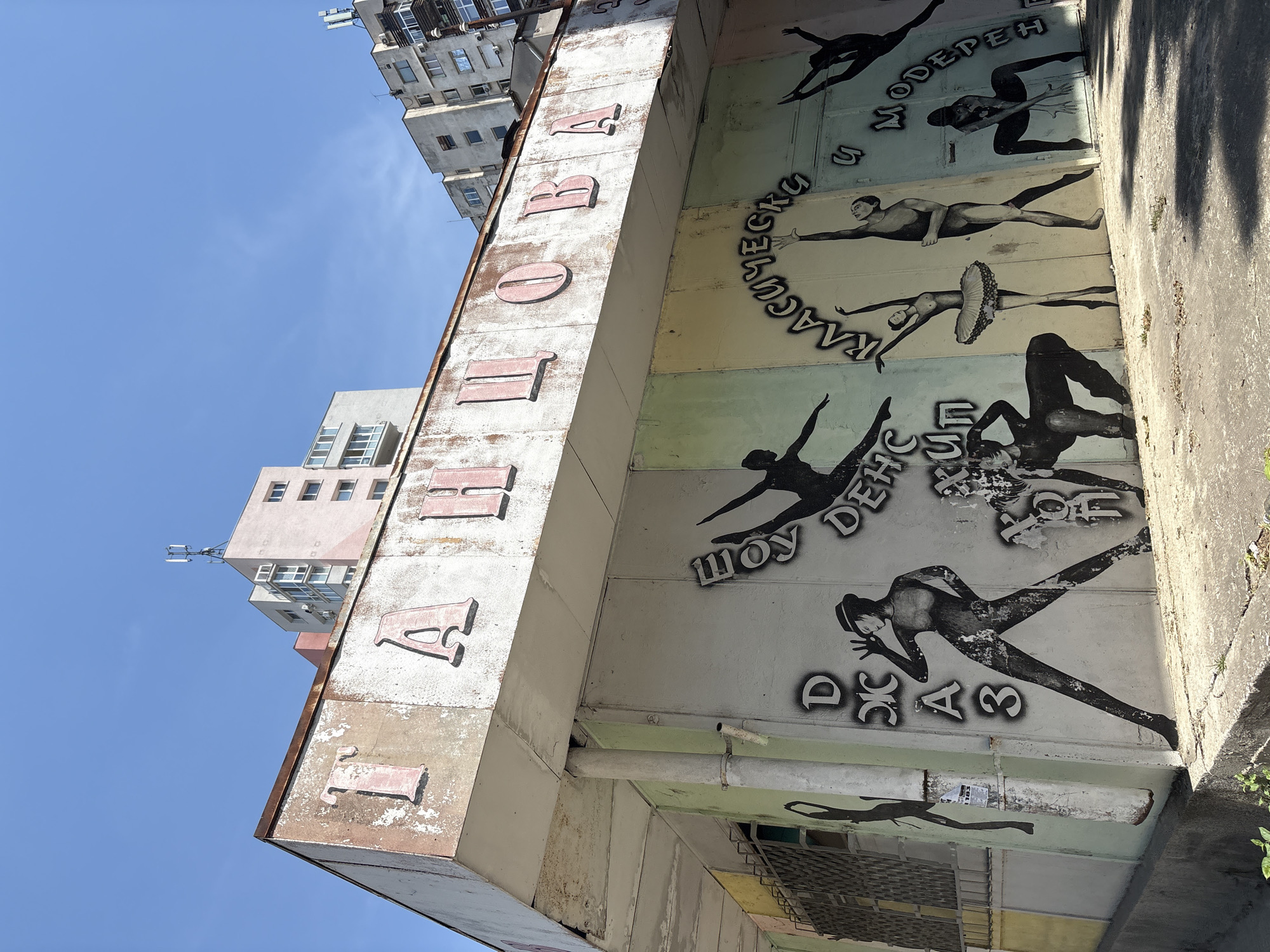The image shows an exterior view of a building in Plovdiv, Bulgaria, likely in a residential or mixed-use area. The building's facade is adorned with faded pink letters spelling out "ТАНЦОВА" (TANTSOVA) across the top, suggesting it might be related to dance. Below the sign, the wall features a mural with stylized black and white figures depicting various dance poses. These figures include individuals in what appears to be ballet attire, as well as more contemporary dance styles. Text in black spray paint is present on the wall, with some legible words like "ДЖАЗ" (JAZZ), "ШОУ" (SHOW), "ДЕНС" (DANCE), "КЛАСИЧЕСКО" (CLASSICAL), and "МОДЕРЕН" (MODERN). The building itself appears aged, with peeling paint and visible rust on the roofline. Behind the building, modern apartment blocks are visible against a clear blue sky. The ground in front of the building is concrete and somewhat weathered, with sparse vegetation. The overall impression is of an older establishment with a vibrant artistic element on its exterior.