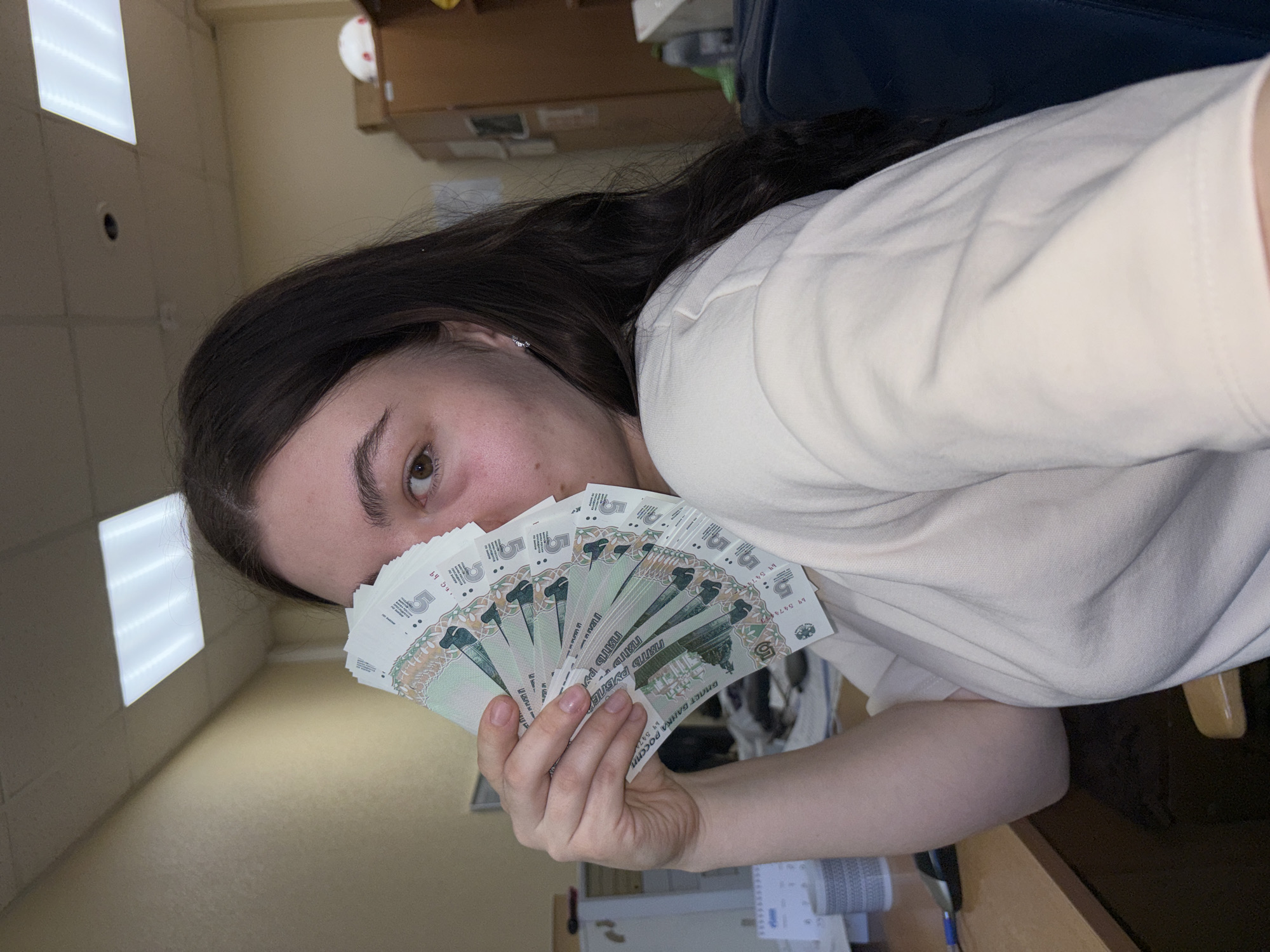 A woman is holding a fan of 5000 Russian Ruble banknotes in front of her face. She has dark hair and is wearing a white shirt. The background appears to be an office setting with fluorescent lighting and cardboard boxes. The banknotes themselves are light green and feature an image of a monument in Perm, Russia, suggesting the photo was taken in that city.