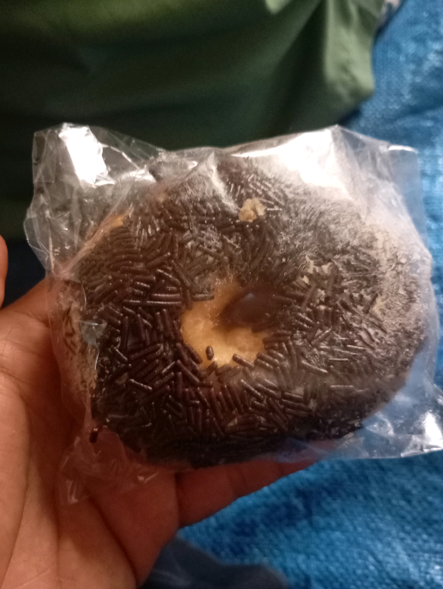 A hand is holding a chocolate donut covered in sprinkles, which is inside a clear plastic wrapper. The donut has a light-colored hole in the center and appears to have some frosting or glaze underneath the sprinkles. The background is out of focus, showing a green fabric surface and a blue textured surface. The image was taken in Sumberjati, Indonesia.