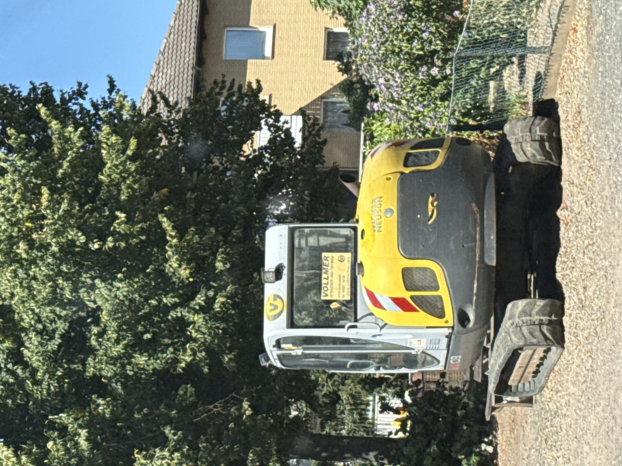 In Bad Sachsa, Germany, a Wacker Neuson mini-excavator is parked on a gravel surface in front of a yellow brick house and lush green trees. The excavator, predominantly yellow and gray, features a cab with glass windows. A yellow sticker with a blue V logo and the text "VOLLMER STRASSEN UND TIEFBAU" is affixed to the rear window of the cab. Another sticker with the Wacker Neuson logo is visible on the front of the machine. The excavator is positioned near a green metal fence and flowering bushes. The sky is clear and blue.