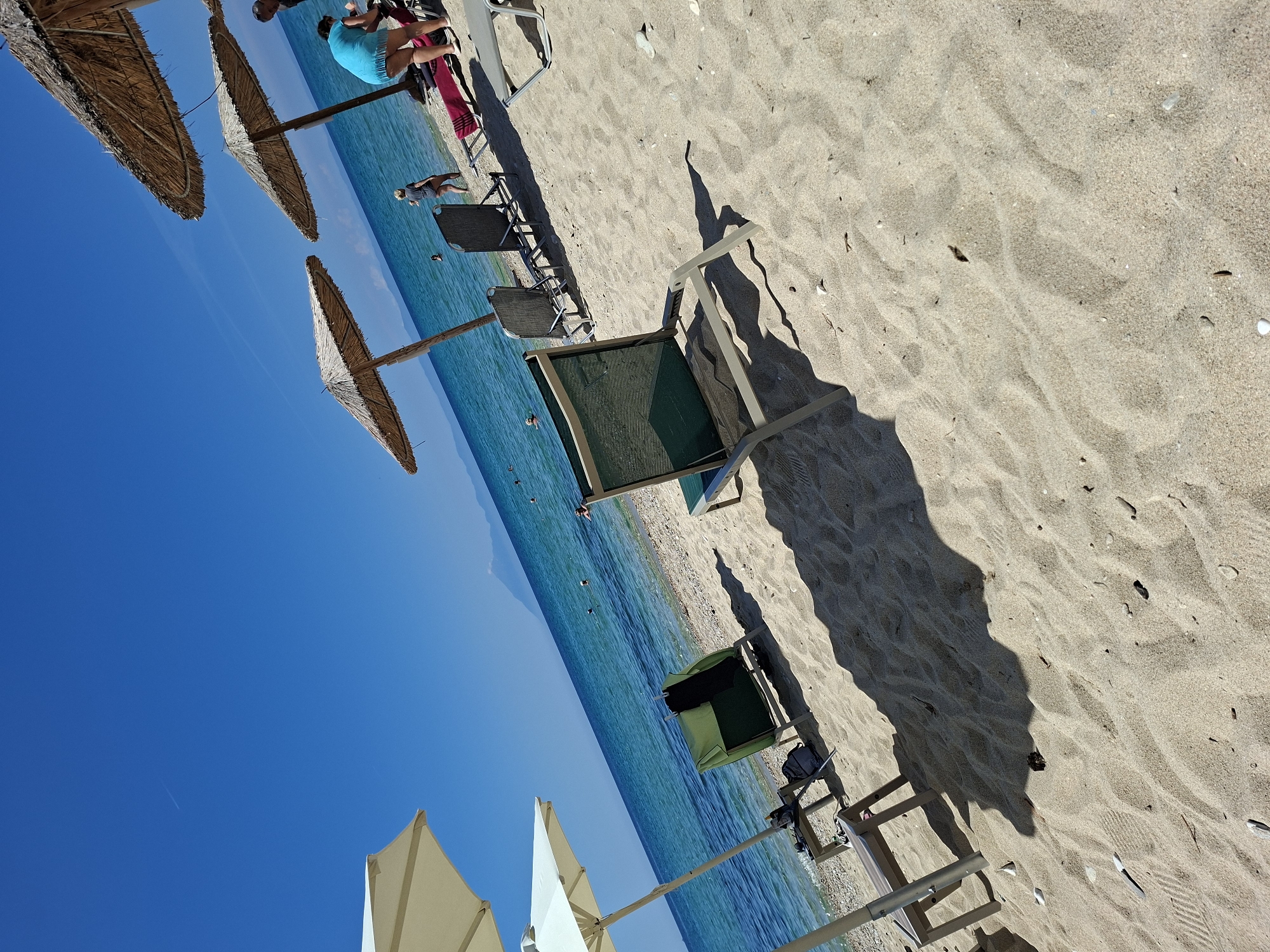 The image depicts a beach scene in Limenaria, Greece. The foreground is dominated by sand with scattered pebbles and debris. Several beach chairs are present, some occupied and some empty. In the middle ground, the turquoise ocean stretches to the horizon, with distant mountains visible. Three thatched umbrellas provide shade, and a few people are seen near the water's edge and in the sea. One person in a blue top and dark shorts is standing on the beach, and another person in dark shorts and a grey top is walking into the water. The sky is a clear, bright blue. The overall impression is a tranquil summer day at the beach.