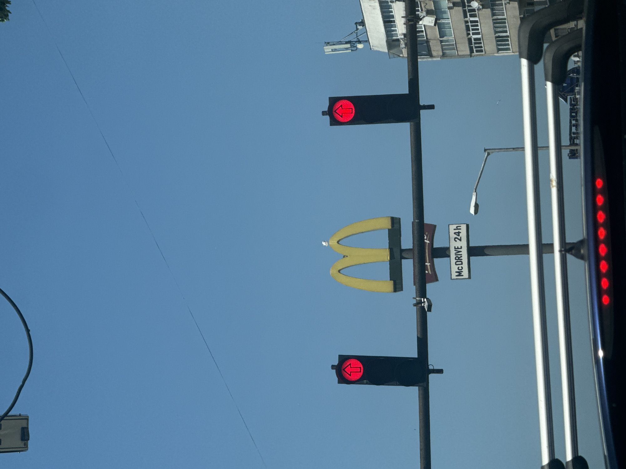 The image captures a street scene in Varna, Bulgaria, under a clear blue sky. Dominating the center is a prominent McDonald's "M" logo, a bright yellow arch. To the left and right of the logo are traffic lights, both displaying a red signal with an upward-pointing arrow. Below the McDonald's sign, a rectangular sign reads "McDRIVE 24h". A street lamp is visible to the right of the sign. In the background, a multi-story building with numerous windows and air conditioning units is situated. The foreground shows the roof and windshield of a vehicle, with brake lights illuminated. A bird is perched atop the McDonald's "M" logo.