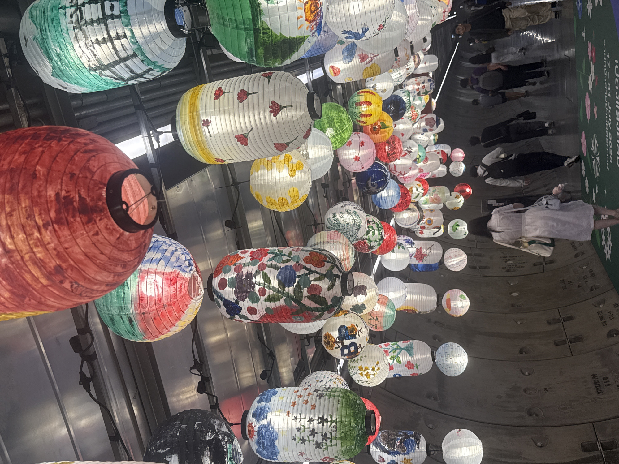 The image depicts an indoor walkway, likely a tunnel or underpass, adorned with numerous hanging paper lanterns of various colors and designs. The lanterns are predominantly cylindrical and spherical, featuring patterns of flowers, landscapes, and some with abstract designs. The walkway walls appear to be made of concrete with visible circular indentations and some markings, suggesting an urban infrastructure setting. The ceiling is metallic, with visible wiring and structural elements supporting the lanterns. Several people are walking through the illuminated space, appearing as silhouettes or partially visible figures from the back and side. They are dressed in casual clothing. The overall atmosphere is festive and decorative due to the vibrant lanterns. This scene is located at the walkway connecting Lumphini MRT station to the adjacent buildings in Bangkok, Thailand.