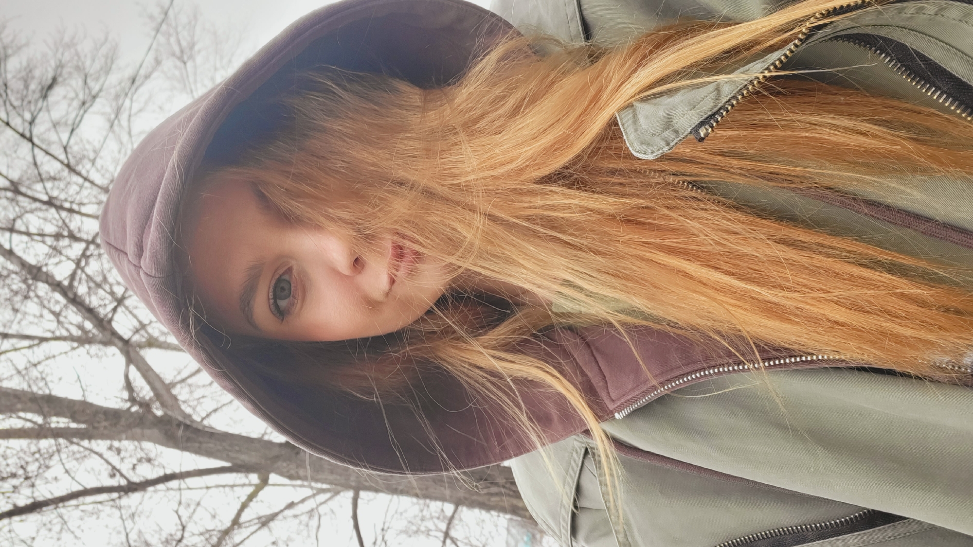 The image, presented in a portrait orientation and rotated 90 degrees counter-clockwise, features a close-up of a person, likely female, with long, light reddish-blonde hair. Her head is framed by the hood of her jacket.
The subject's face is partially obscured by her flowing hair. One light-colored eye (green or hazel) is visible, looking towards the camera. Her nose and part of her mouth, with a subtle, closed-lip expression, are also visible. Her long, straight hair cascades across her face and extends out from under her hood and jacket.
She is wearing a hooded jacket. The outer layer appears to be olive green or muted khaki, with prominent zippers visible on the chest area. The inner lining and hood are a contrasting dark, muted purple or maroon color.
The scene is set outdoors against a light, overcast sky. In the background, numerous bare tree branches are visible, suggesting a colder season like late autumn, winter, or early spring in Chaikovsky, Russia. The overall impression is one of chilly weather, consistent with the heavy hooded jacket.
There is no discernible activity beyond the subject looking at the camera. Her expression is neutral, perhaps serene. No visible text is present in the image.