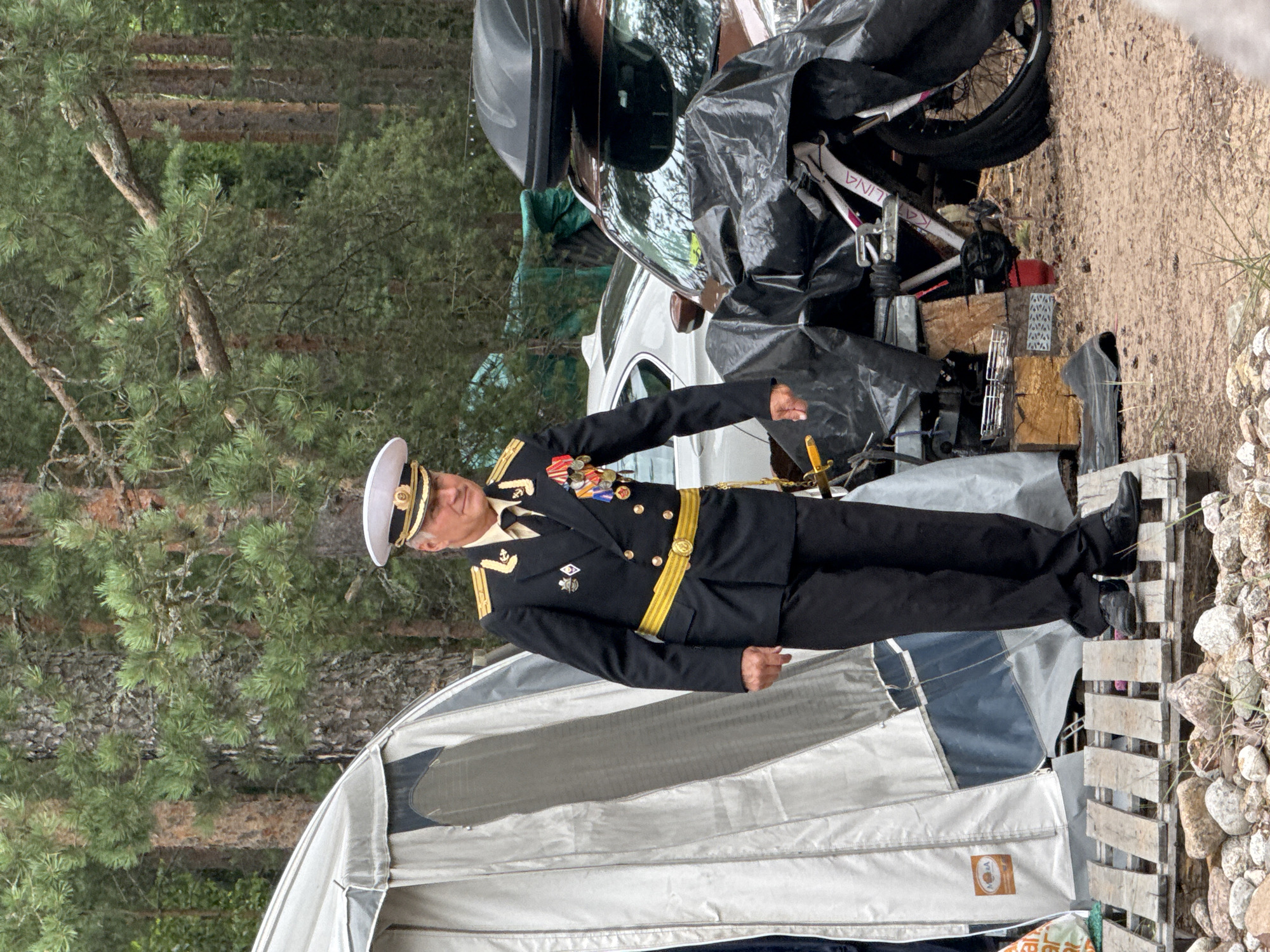 The image shows a man in a naval uniform standing on a wooden platform next to a tent. He is wearing a white hat with a gold emblem, a dark blue double-breasted jacket with gold trim and epaulets, and black pants. He has several medals pinned to his chest. Behind him are trees, a white car, a brown car with a roof-top carrier, and a bicycle covered with a black tarp. The ground appears to be sand and gravel. The scene is set outdoors, likely in a campground. The image does not provide specific information about the location within Zelenaya Roshcha, Russia, but the general environment suggests a natural setting.