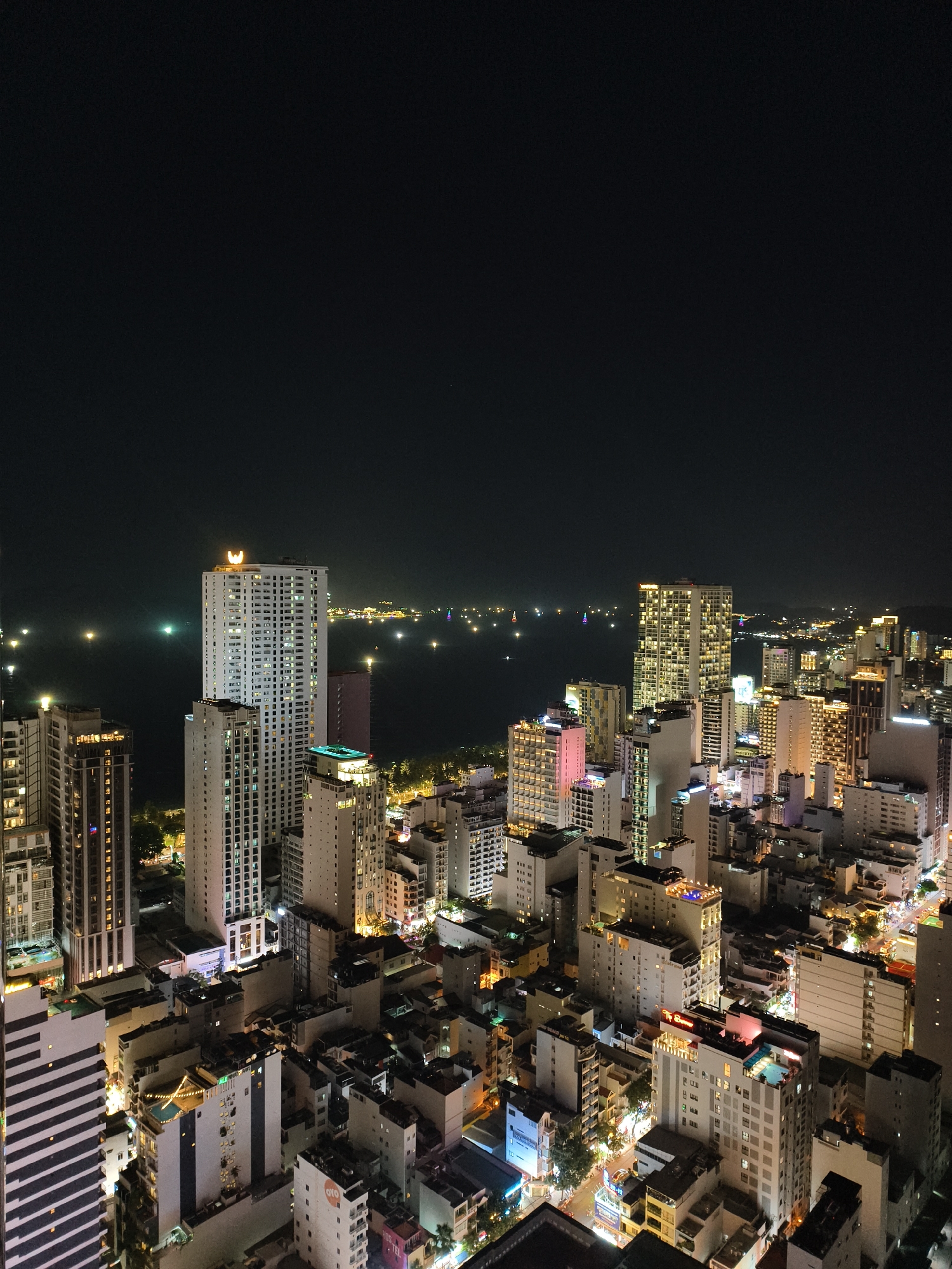 This high-angle, wide shot captures a dense coastal city at night, presenting a sprawling urban landscape illuminated by countless lights. The scene is dominated by numerous high-rise buildings and skyscrapers, with their windows glowing in various shades of yellow, white, pink, and blue, creating a vibrant tapestry against the dark night sky.
In the mid-ground, a large body of water, likely an ocean or bay, stretches into the distance, dotted with numerous bright lights from what appear to be boats or distant shorelines. The tallest and most prominent building in the center-left is a white skyscraper with a distinctive, glowing crescent moon or stylized 'W' shaped logo at its very top.
The city's density is evident, with buildings of varying heights clustered closely together. Several buildings feature brightly lit rooftop pools. Visible text on signs includes "OYO" on multiple buildings, "HOS TEL" on a building towards the bottom-right, and partial sightings of "THE D", "Luxury Apartment", "MUONG T", and "NOVOTEL". The clear night sky is completely dark, devoid of stars or clouds, emphasizing the artificial glow of the urban environment. No people or specific activities are discernible, giving the impression of a static, majestic cityscape after dusk.