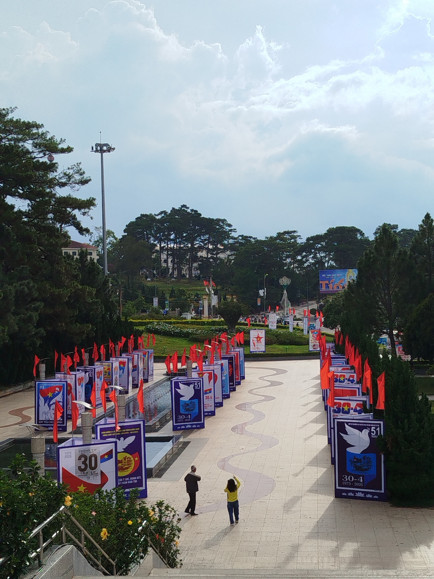 The image shows a public square in Da Lat, Vietnam, adorned with political banners and flags. These banners, arranged along a wavy pedestrian walkway, commemorate historical events, notably the "30-4," likely referring to April 30th, the anniversary of the Reunification Day of Vietnam. Two individuals are present in the scene: one man in dark attire and a woman in a yellow shirt, walking along the walkway. The surrounding environment includes lush greenery, pine trees, and a view of buildings and a light post in the distance. The sky is partially cloudy.