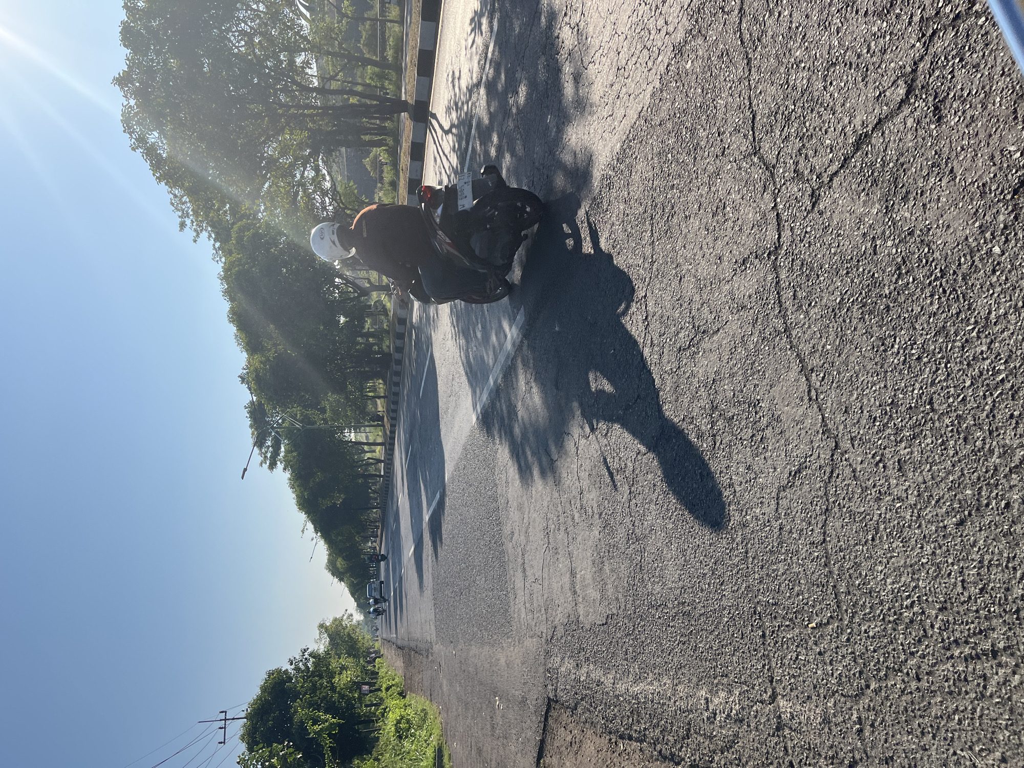 The image captures a sun-drenched road in Siring, Indonesia, with a clear blue sky overhead and bright sunlight casting long shadows. The road surface is visibly cracked and uneven, with patches of newer asphalt laid over older, damaged sections. Lush green trees line the right side of the road, creating a canopy that partially shades the pavement. On the left, dense foliage and utility poles are visible.
In the foreground, a person wearing a white helmet and a maroon shirt rides a dark-colored motorcycle away from the camera, casting a prominent shadow on the road. The motorcycle has a license plate that is partially obscured. Further down the road, other vehicles, including cars and motorcycles, are visible in the distance. The overall scene depicts a typical day on a well-trafficked, albeit somewhat deteriorated, road in a tropical urban or suburban environment.