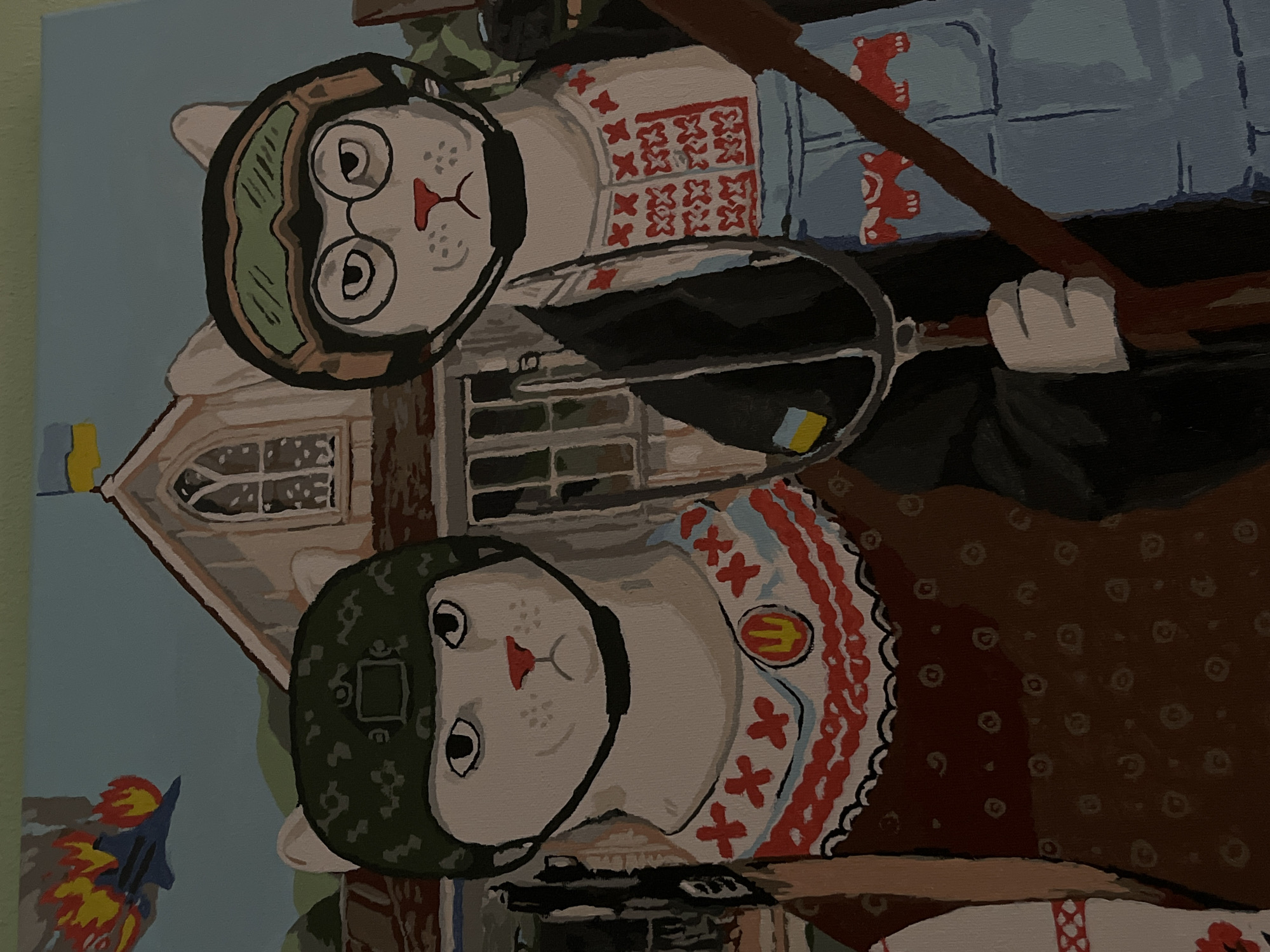 This is a painting depicting two anthropomorphic cats in what appears to be a stylized, possibly surreal, outdoor scene. Both cats are wearing military-style helmets. The cat in the foreground, on the right, is wearing a green helmet with a camouflage pattern and a white ruff collar with red embroidered crosses and a yellow emblem. The cat in the background, on the left, is wearing a similar green helmet with a different pattern and round glasses. This cat is also wearing a white shirt with red embroidered patterns. They are positioned in front of a building that resembles a church or a historical structure with arched windows.
The scene is rendered in a folk art style with bold colors and simplified forms. The background is a pale blue sky. To the left of the frame, a blue rocket or projectile with flames is depicted. The overall tone of the artwork could be interpreted as patriotic or symbolic, especially given the context of Lviv, Ukraine. The cats' expressions are neutral or slightly solemn. There is no visible text in the image. The time of day and weather are not explicitly indicated, but the lighting suggests daylight.