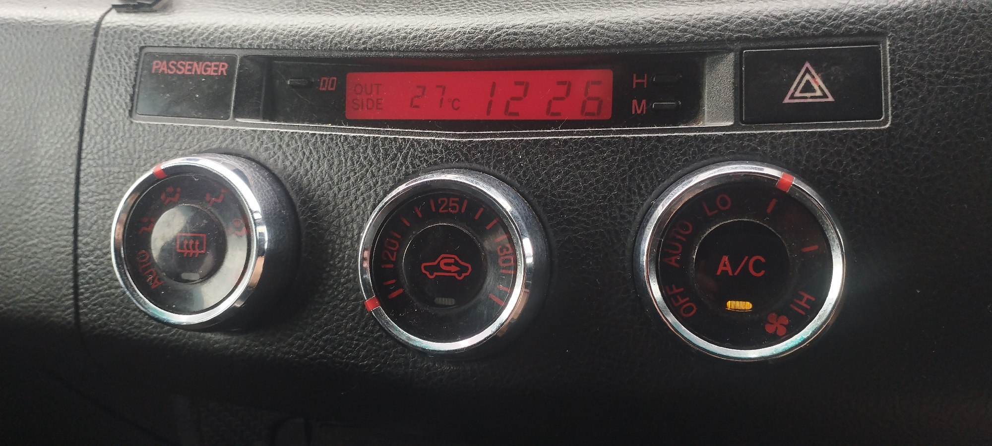 The image shows a close-up of a car's climate control panel. The panel has a digital display showing "OUT SIDE 27°C 12:26". There are three circular knobs below the display. The left knob has symbols for defrosting and fan speed. The middle knob has symbols for airflow direction. The right knob controls the A/C and fan speed, with "LO", "AUTO", "HI", and an A/C indicator light illuminated. To the right of the digital display is a hazard light button with a red triangle symbol. The panel is made of black, textured plastic. There are no people or animals visible in the image. The environment appears to be the interior of a car. There is no information available to determine the location of Покосное, Russia in relation to this image.