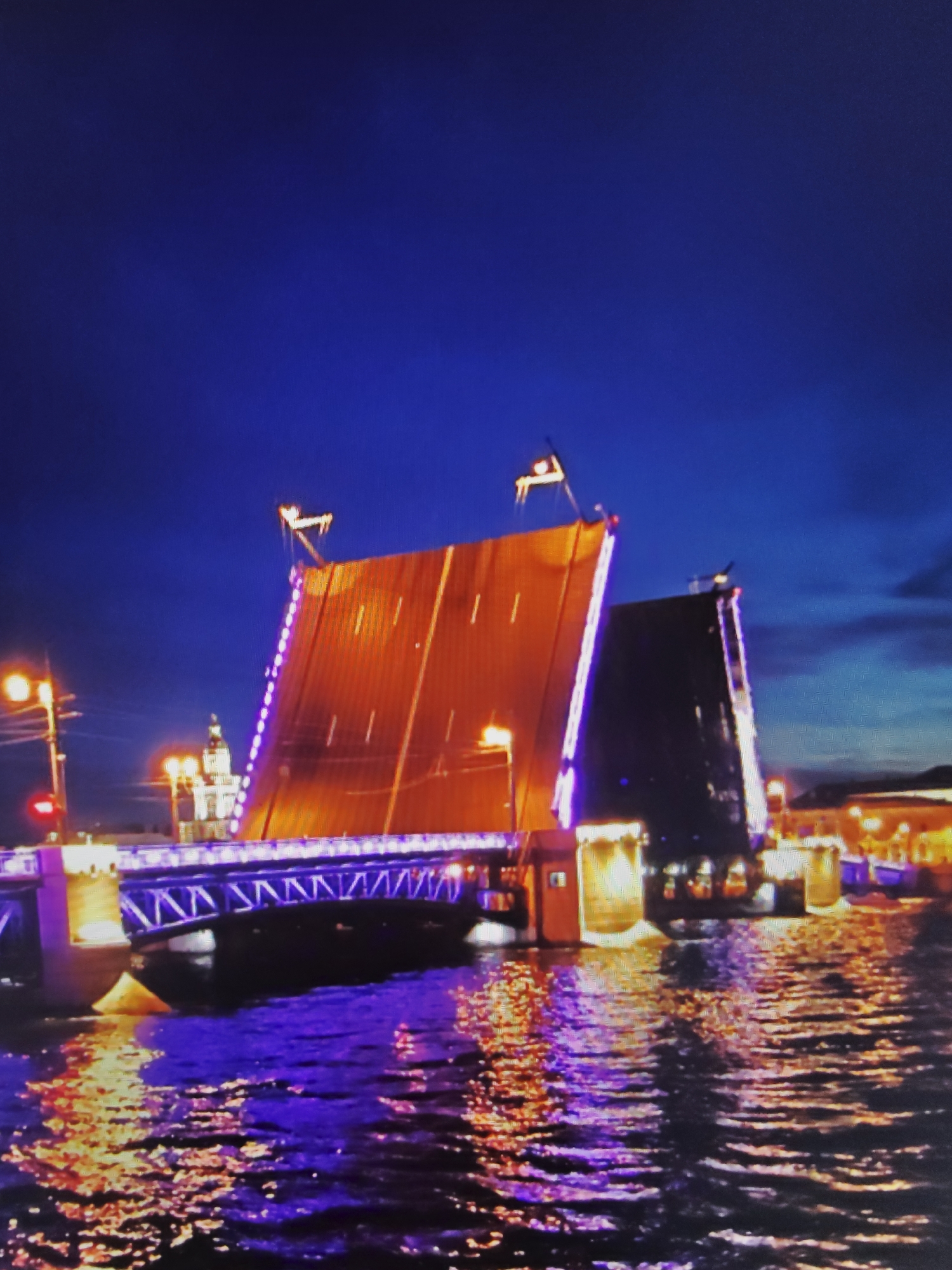An image captured at night shows a large, illuminated drawbridge in St. Petersburg, Russia. The bridge is in its raised position, with two sections angled upwards, creating an imposing silhouette against the dark blue sky. The bridge is adorned with strings of purple lights along its edges, which cast a vibrant glow on the structure and the water below.
The water in the foreground reflects the colorful lights from the bridge and the surrounding cityscape, creating a shimmering, artistic effect. The reflections are a mix of purples, oranges, and yellows, broken by the ripples of the water.
In the background, city lights are visible, suggesting a bustling urban environment. A dome-shaped building, likely a church or a significant landmark, is illuminated and stands out against the night sky. The overall scene is one of architectural grandeur and nighttime vibrancy, with the drawbridge as the central focal point. There are no people or animals visible in the frame.