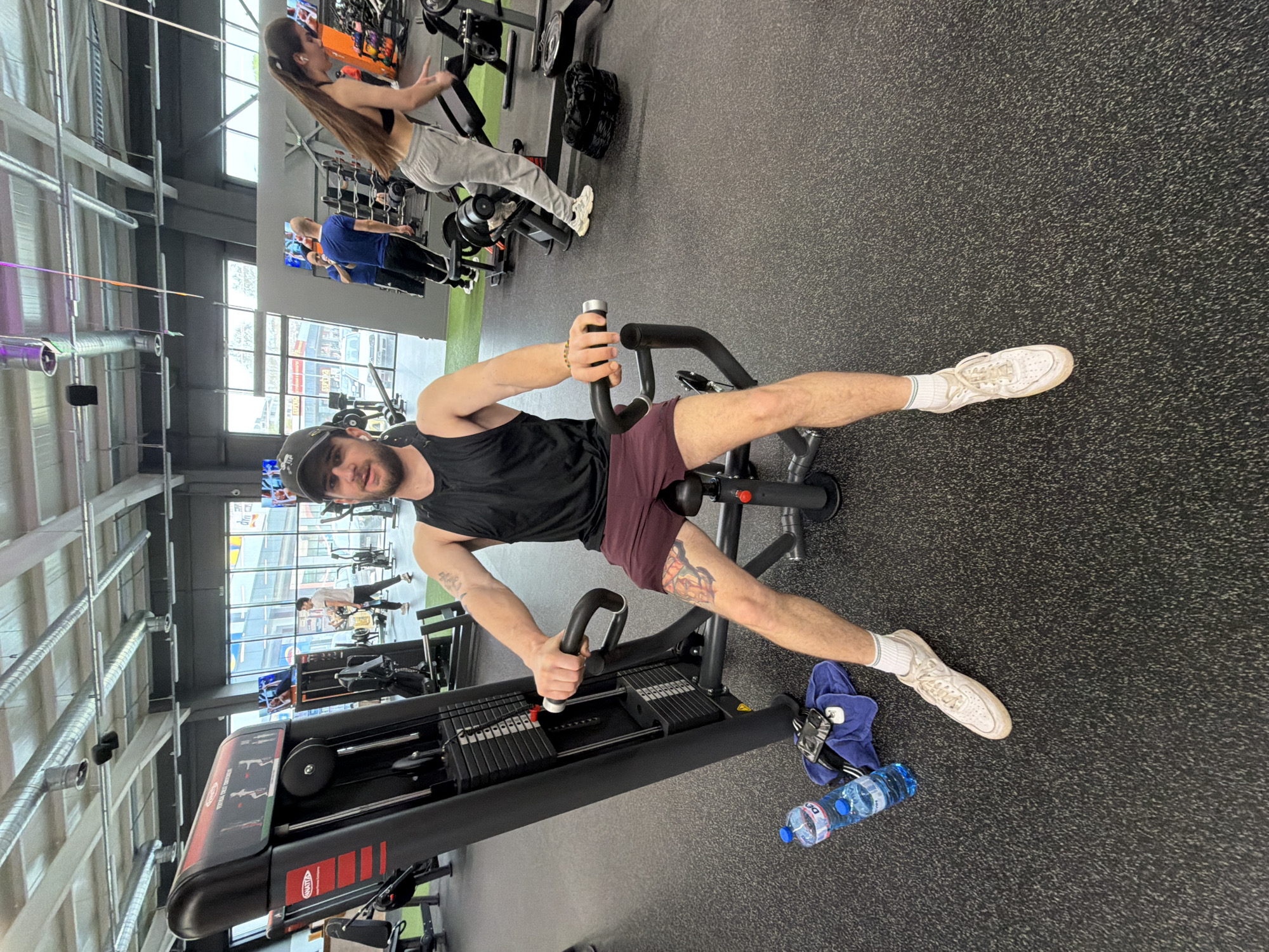 The image shows a man in a gym, likely in Plovdiv, Bulgaria, given the location tag. He is sitting on a seated ab crunch machine, wearing a black tank top, maroon shorts, a baseball cap, and white sneakers. He is grasping the machine's handles, seemingly mid-exercise. A tattoo is visible on his right thigh. Next to the machine, on the floor, are two water bottles and a blue towel. Other gym-goers are present in the background, using different exercise equipment. The gym has large windows and an industrial-style ceiling with visible ductwork.