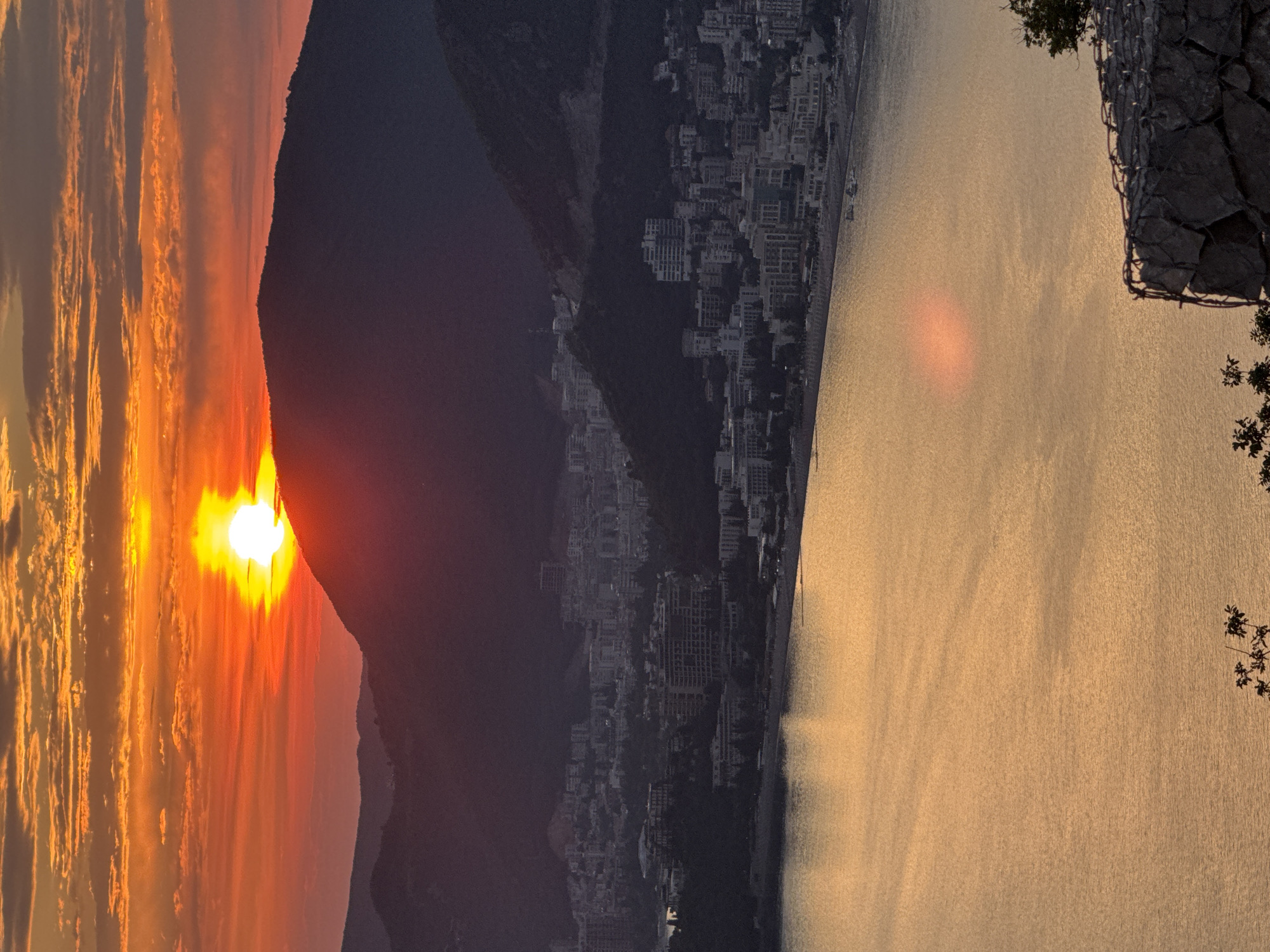 The image depicts a sunset scene overlooking a coastal city in Čelobrdo, Montenegro. The sun, a bright orange orb, is low on the horizon, casting a warm, golden glow across the sky and reflecting on the calm water. The sky is filled with streaks of orange and pink clouds, suggesting fair weather.
In the foreground, a rocky structure with wire mesh is visible on the right, partially obscuring the view. Silhouetted against the vibrant sky, a mountain range forms a dark backdrop to the scene. Nestled on the slopes of the mountains is a dense urban landscape, characterized by numerous buildings and structures that are visible even in the dimming light. The city appears to be situated along the coastline, with the water stretching out towards the horizon.
The overall mood of the image is serene and peaceful, capturing the tranquil beauty of a sunset in a picturesque coastal setting. The specific location cues of Čelobrdo, Montenegro, combined with the visual elements, evoke a sense of natural beauty and urban integration. There is no visible text in the image.