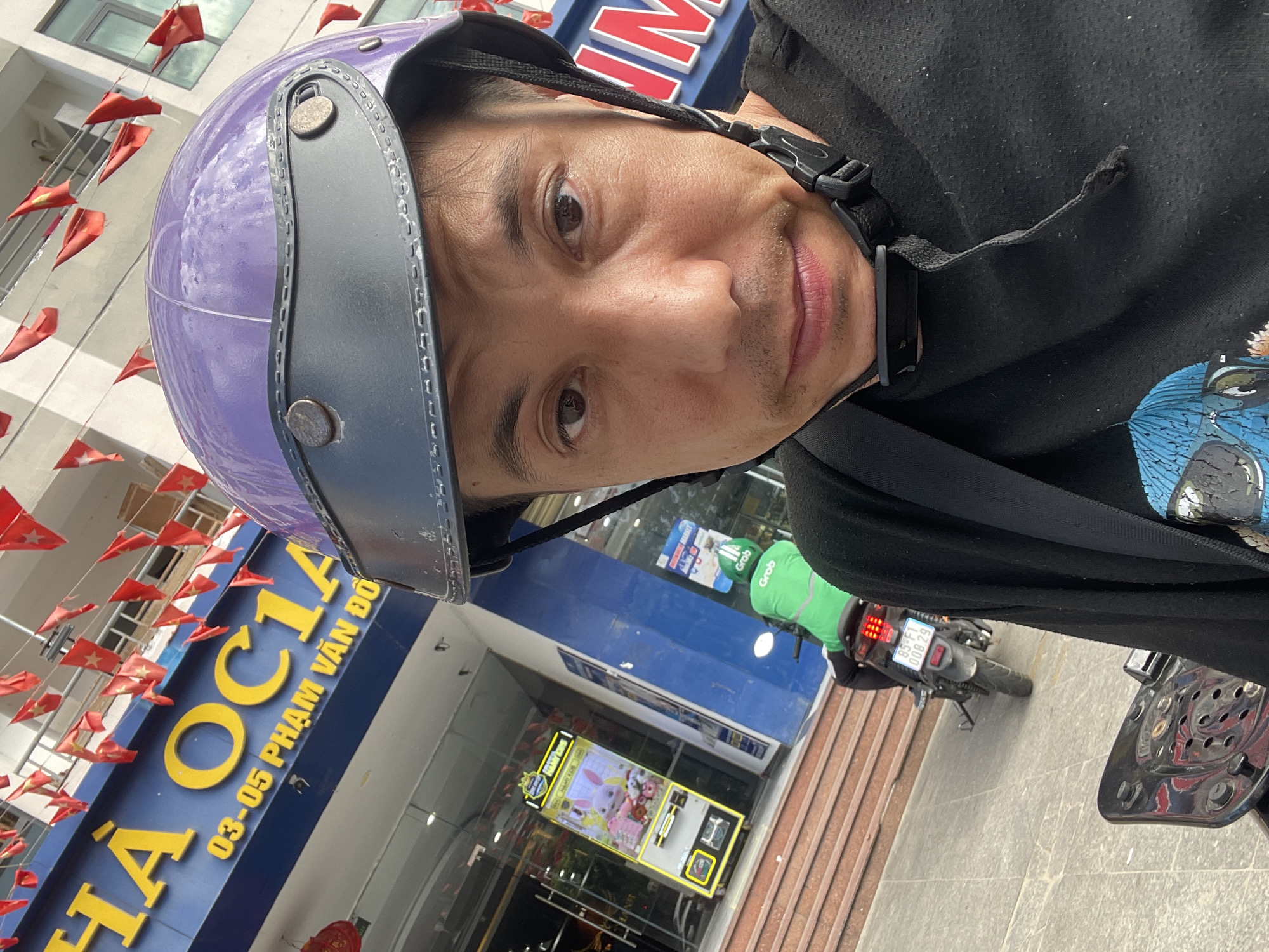 This media file is a selfie taken by a person wearing a purple helmet and a black shirt. The person's face is close to the camera, and they appear to be looking directly at it with a neutral expression. They have dark hair and a light complexion, with a hint of stubble on their chin.
The background of the image suggests an outdoor setting in Vietnam, with a building displaying a large blue and yellow sign that reads "HA OCIA 03-05 PHẠM VĂN ĐỒ". Red Vietnamese flags are strung along the facade of the building, adding a festive touch.
In the middle ground, a claw machine game is visible through the glass storefront, displaying plush toys. Further in the background, a motorcycle is parked, with a person in a green Grab uniform on it. The license plate of the motorcycle is partially visible, reading "85-F1 008.29".
The overall impression is a candid snapshot of everyday life in a Vietnamese urban environment, possibly taken during daylight hours. The combination of the street scene, the signage, and the presence of a delivery rider suggests a commercial or busy area.