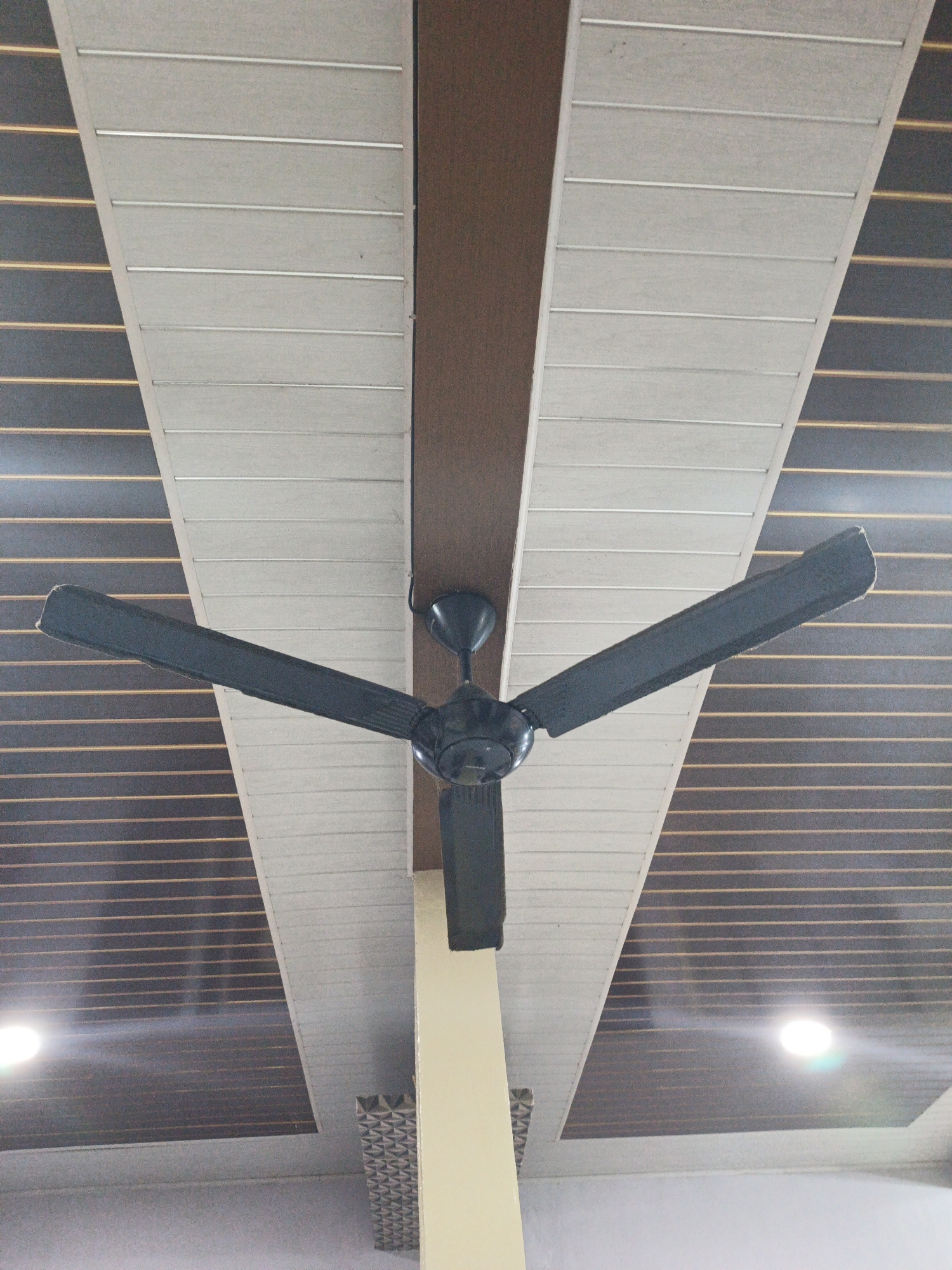 A low-angle shot captures a black ceiling fan mounted on a brown wooden beam against a backdrop of a sloped ceiling composed of white and dark brown paneled sections. The white panels are horizontally oriented and separated by thin silver strips, while the dark brown panels feature multiple thin, evenly spaced horizontal yellow lines. Two circular lights are visible, one on the left and one on the right side of the ceiling. The fan's blades are extended, suggesting it might be in use or recently stopped. The location is identified as Kelapapati, Indonesia.