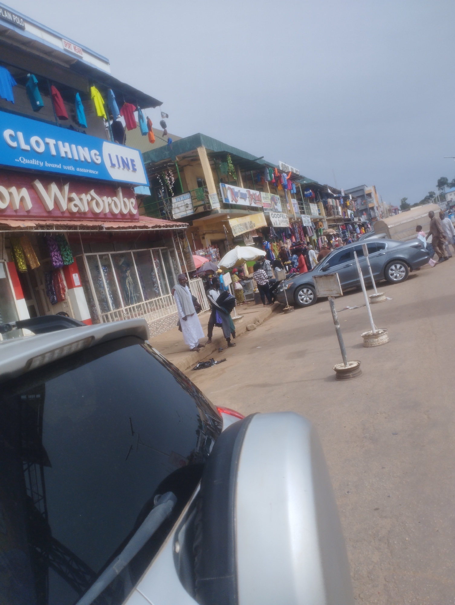 The image captures a street scene in Jos, Nigeria, likely a commercial area characterized by shops and pedestrian activity. In the foreground, the side mirror and partial view of a silver car are visible, suggesting the photograph was taken from within or near a vehicle. The car's windshield reflects the sky and parts of the opposite storefronts.
Dominating the mid-ground are a row of shops, one prominently displaying a large blue sign that reads "CLOTHING LINE" with the tagline "...Quality brand with assurance." Below this sign, another shopfront features a red awning with the name "on Wardrobe" in white, stylized lettering. Various colorful garments are displayed on racks and in shop windows.
Beyond these shops, a two-story building with several businesses on its facade is visible. Signs for "GODFEL," "NEW IMAGE," and other retail outlets can be deciphered. Several people are present on the street. Some are walking, while others appear to be customers or shopkeepers. Two individuals are standing near the "on Wardrobe" shop, one wearing a white robe and head covering. A gray sedan is parked on the street, and several metal posts with circular bases are interspersed along the roadside.
The environment appears to be a busy urban street with a mix of commercial and pedestrian activity. The sky is overcast, indicating a cloudy day. The ground surface is paved, with some dirt and debris. The overall impression is that of a bustling marketplace or a commercial street in a Nigerian city.