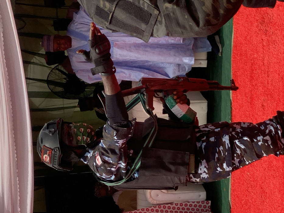 The image depicts a security operative and civilians at an event in Ifira, Nigeria. In the foreground, a dark-skinned male security operative is positioned facing slightly right, extending his right arm and pointing forward with a gloved index finger. He wears a dark tactical helmet with a red and white patch and an additional dark patch, an olive green/khaki patterned face mask with small red motifs, a blue/black/white digital camouflage uniform, and a dark tactical vest. A green sling diagonally crosses his chest. He carries an AK-pattern rifle with reddish-brown furniture, its magazine wrapped in green, white, and green material. The operative stands on a red carpet.
In the mid-ground, a man wearing a light blue traditional gown and cap stands, looking towards the left with a slight smile. Behind him and to his right, the shoulder and upper arm of another individual in a dark green/camouflage uniform are partially visible. To the far left background, blurred figures in light-colored attire are seated on a white upholstered bench. A large black industrial fan is visible in the background, to the right of the seated figures. The scene is set under a light-colored canopy or tent structure.